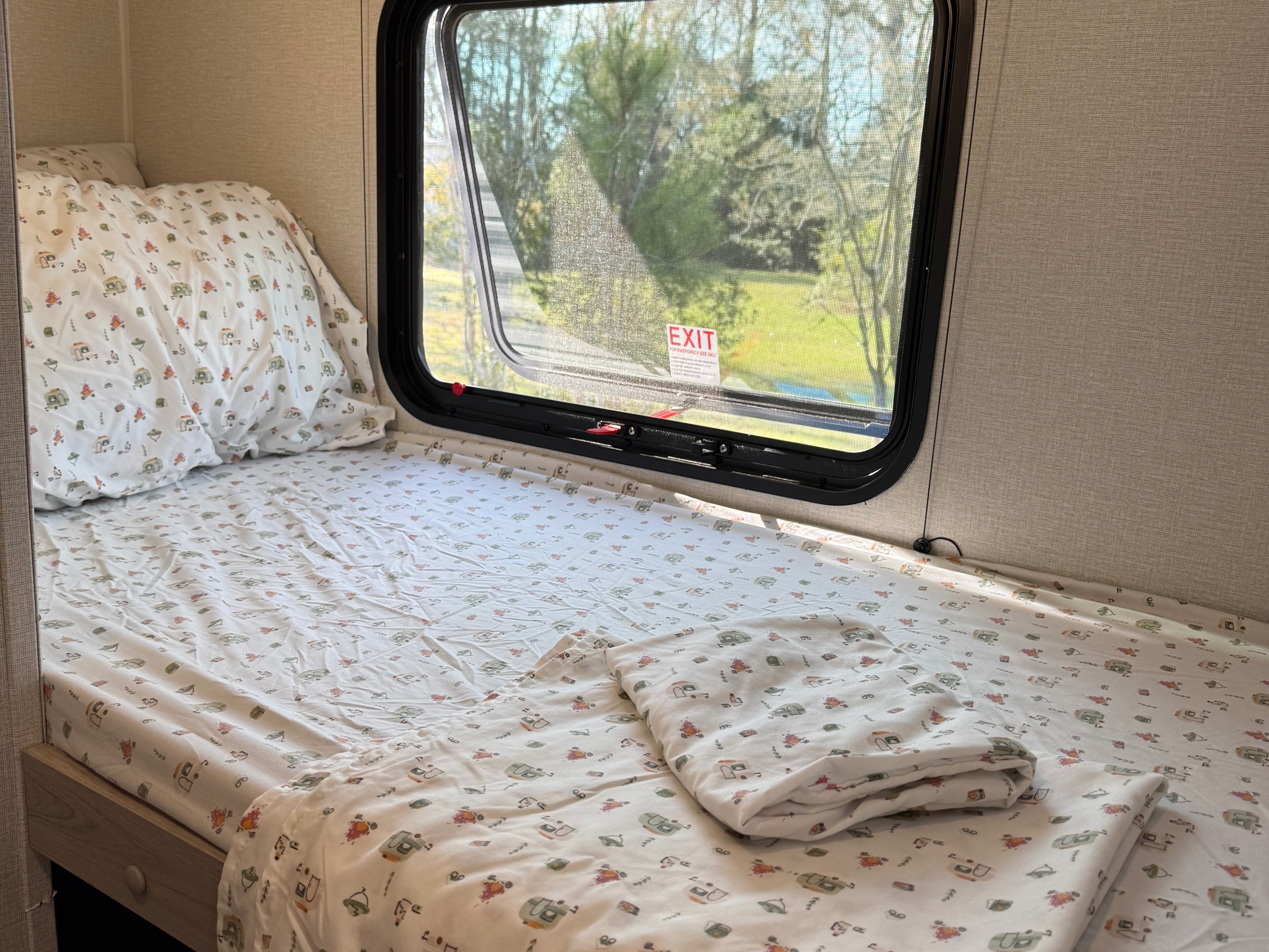Cozy RV rental bunk bed with camping-themed sheets, a window overlooking lush trees, and natural light.. Heartland RVs Pioneer 2025