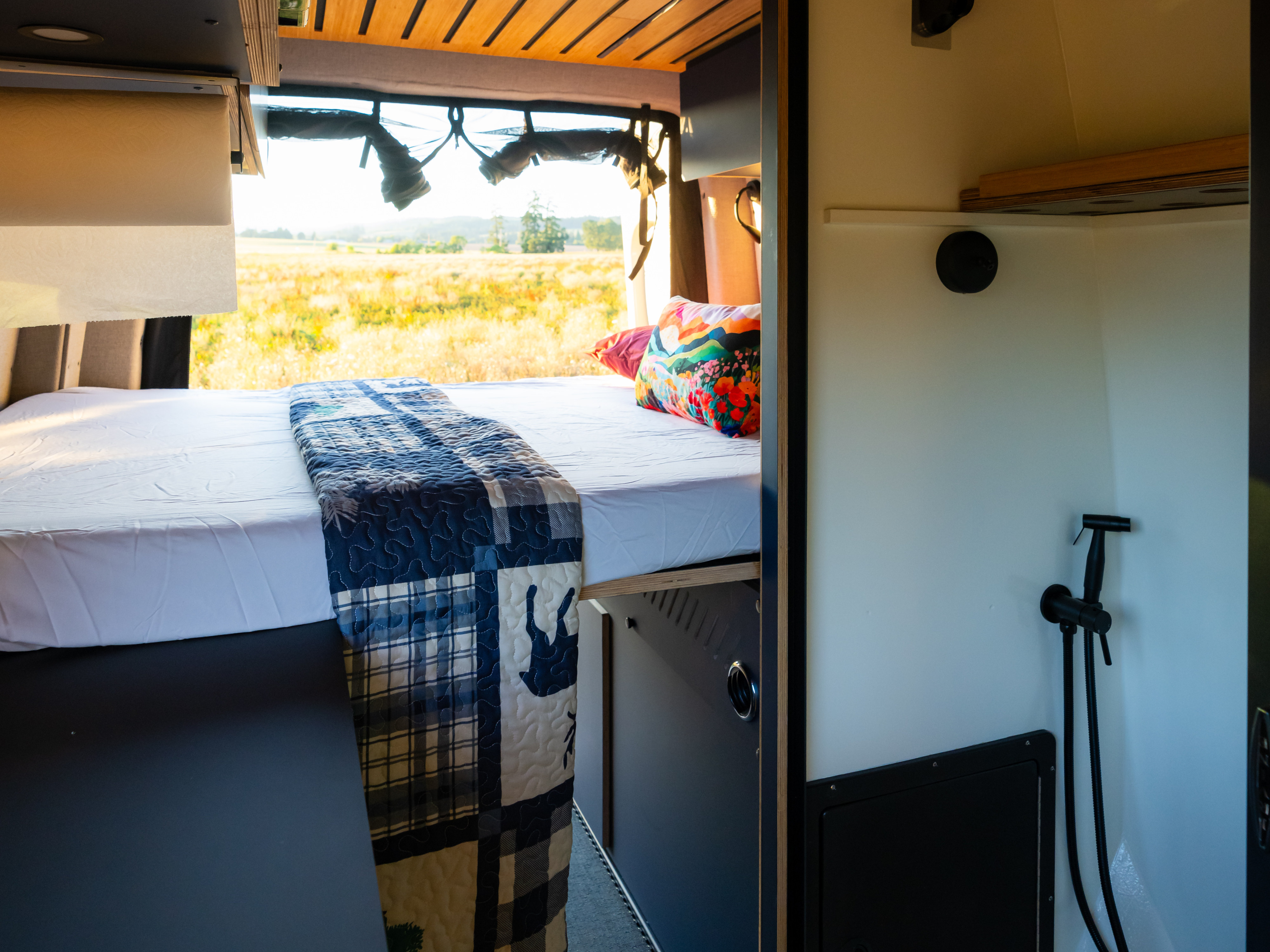 This RV rental's interior features a comfy bed with a blue patchwork quilt, vibrant pillow, and a sunny field view. Shower visible.. Mercedes Sprinter 2026