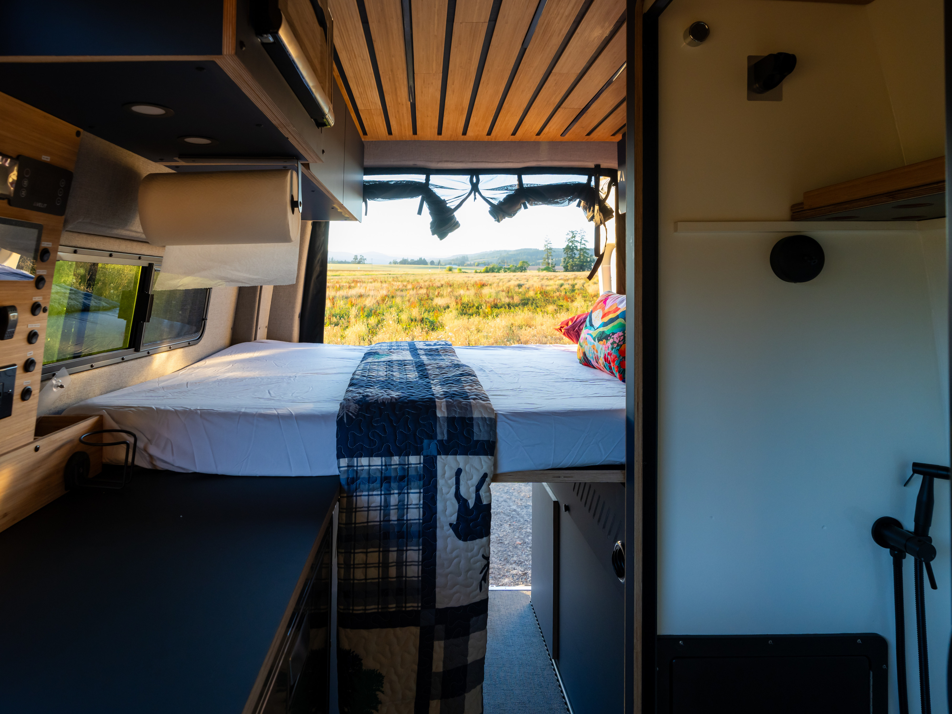 Wood-paneled RV rental interior featuring a cozy bed, controls, and a scenic field view from the open van rear.. Mercedes Sprinter 2026