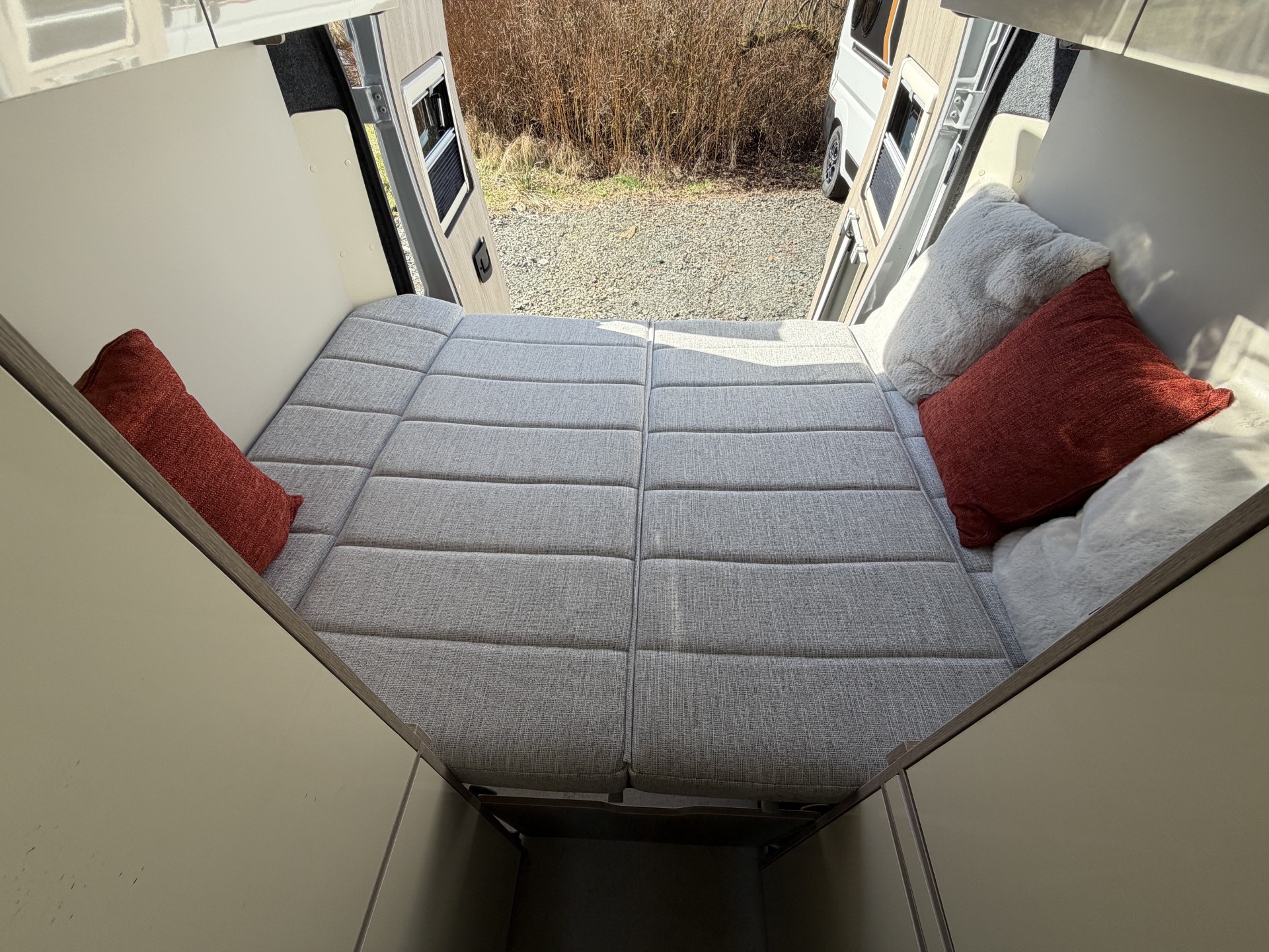 Inside this inviting RV rental: a plush grey bed with red/white pillows, facing open back doors revealing a gravel outdoor scene.. WildAx Aurora XL 2026