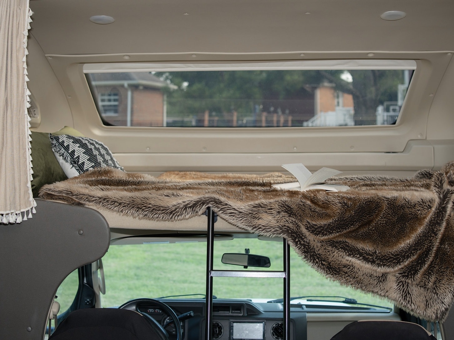 Cozy RV rental bunk bed with faux fur blanket, open book, and ladder. Through the windshield, a green landscape is visible.. Entegra Coach Odyssey 2020
