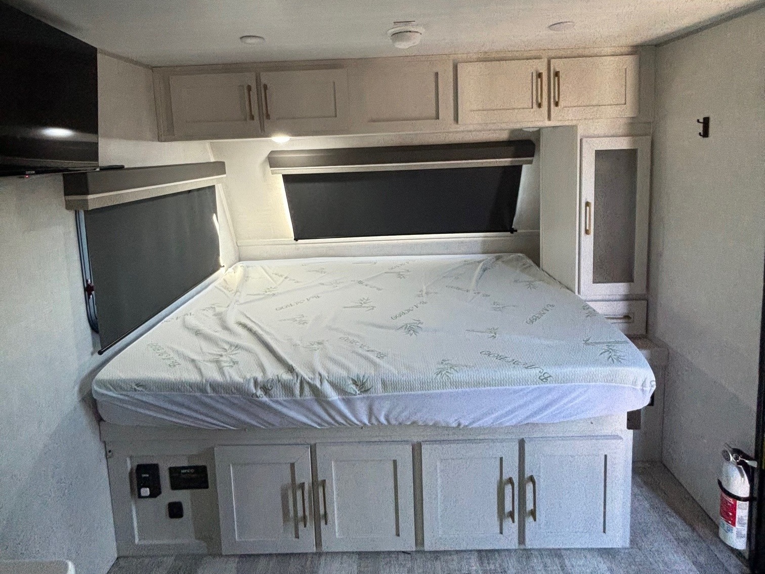 This RV rental bedroom features a comfortable bed, ample storage cabinets above and below, dark window blinds, and a TV.. No Boundaries 18 2025