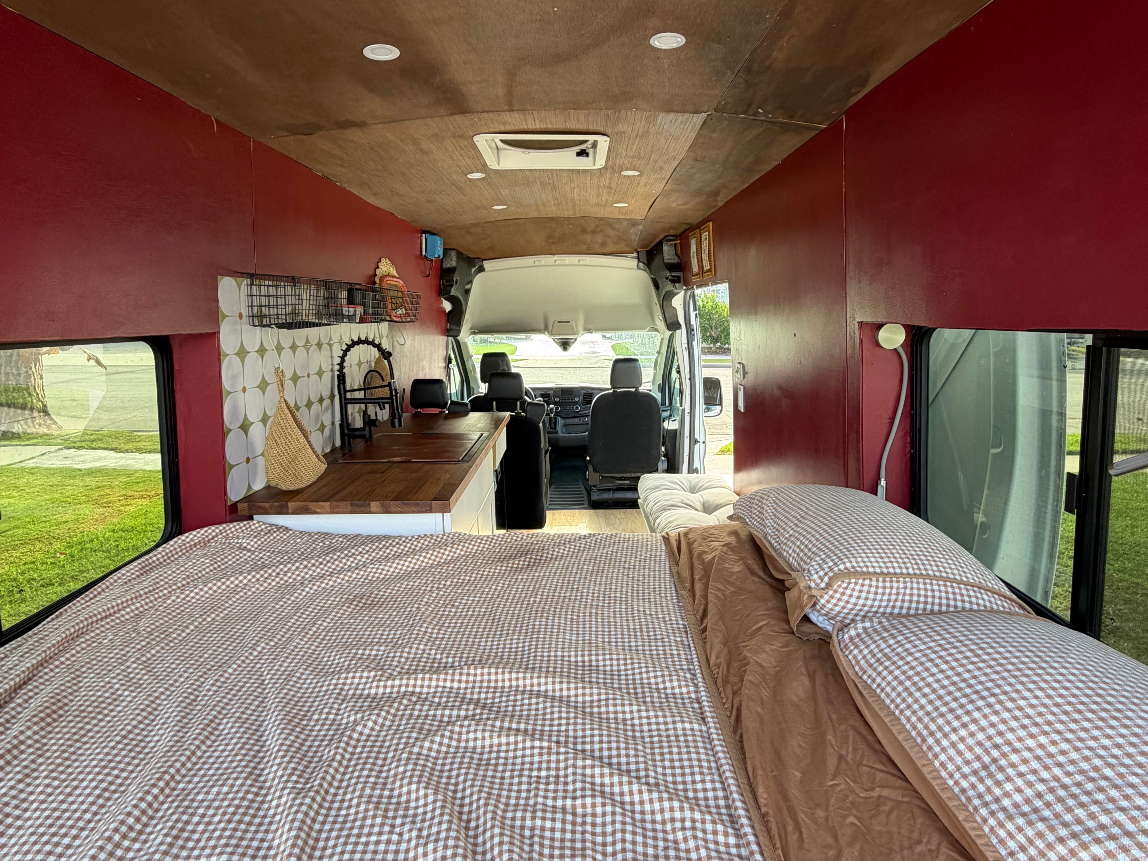 Cozy RV rental interior: a bed with brown gingham bedding, kitchen area, red walls, wooden ceiling, and front van seats.. Ford Transit 2021