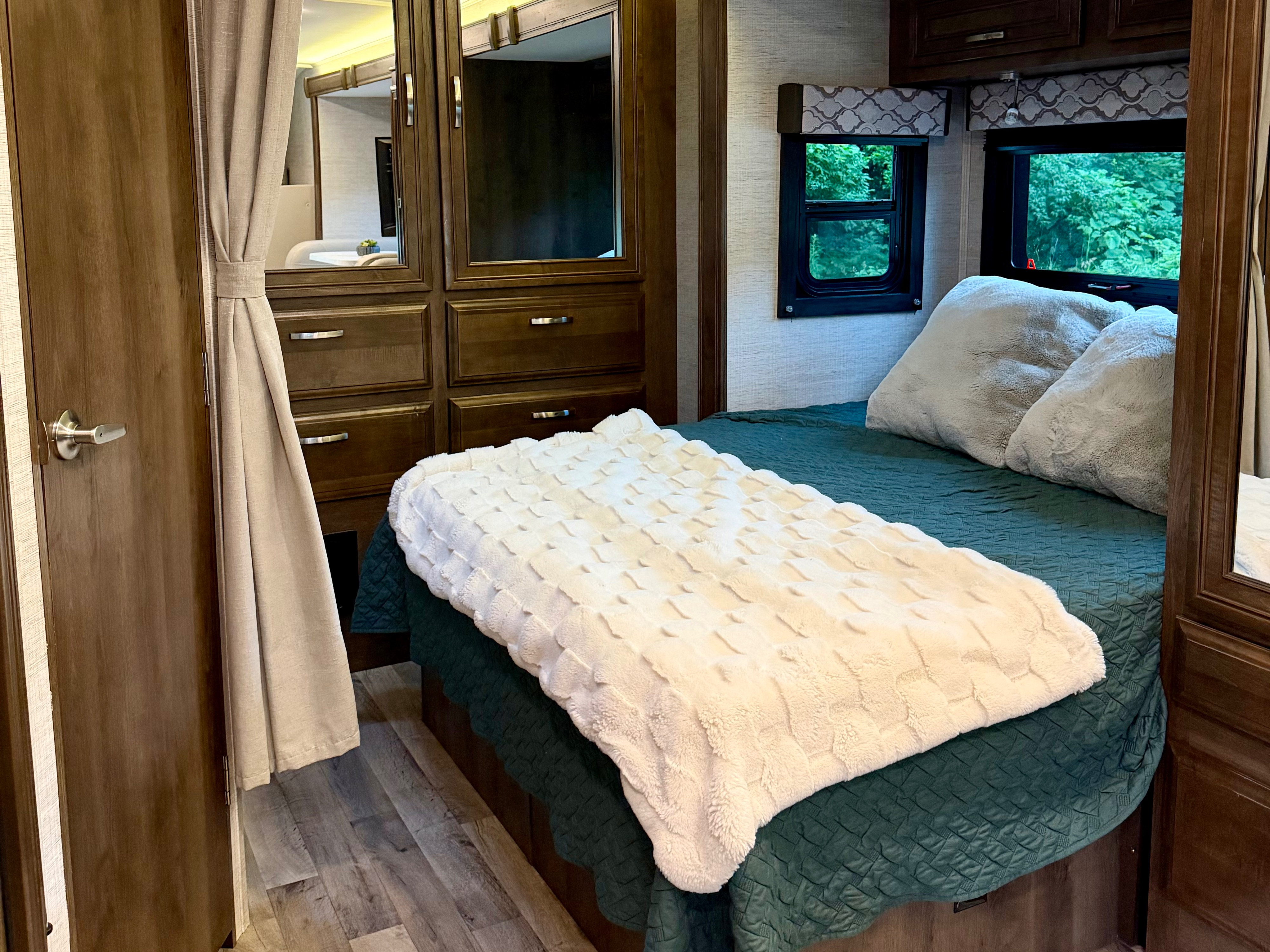 Cozy RV rental bedroom featuring a bed with teal bedding, a fluffy white blanket, wood cabinets, and windows with green views.. Jayco Redhawk 2022