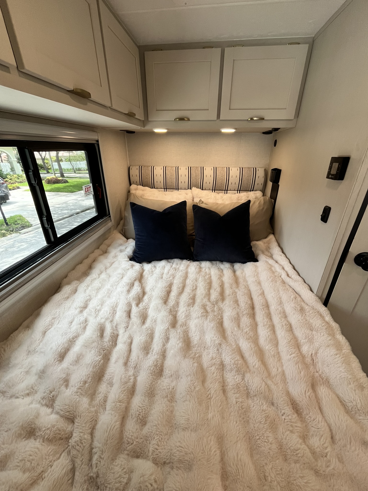 Relax in this inviting RV rental bedroom featuring a plush bed, blue pillows, overhead cabinets, and a scenic window view.. Forest River Coachmen Prism Elite 2026