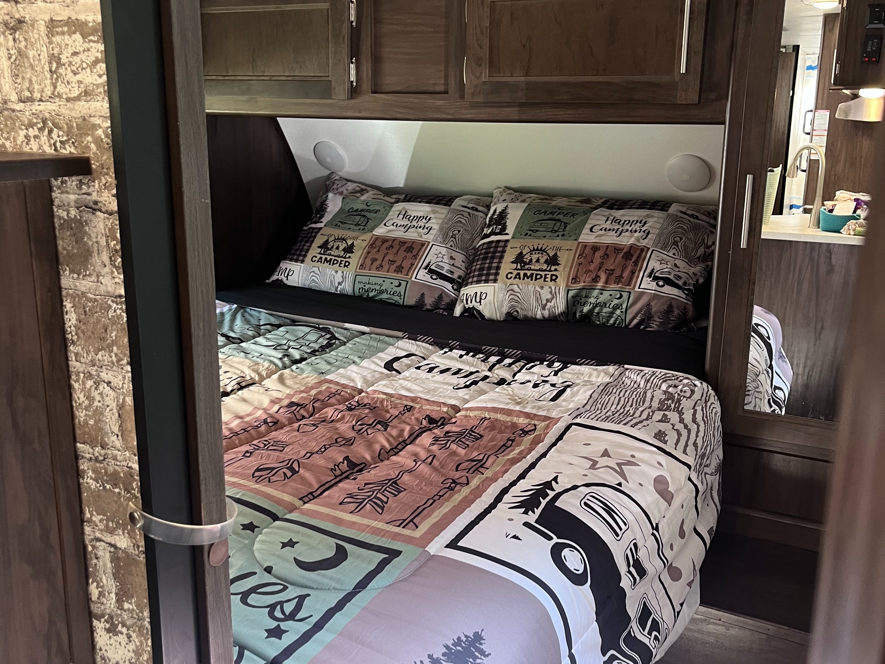 Cozy RV rental sleeping area: bed with "Happy Camping" pillows and a camping-themed comforter. Wooden cabinets and kitchen reflection.. Forest River Cherokee Grey Wolf 2018