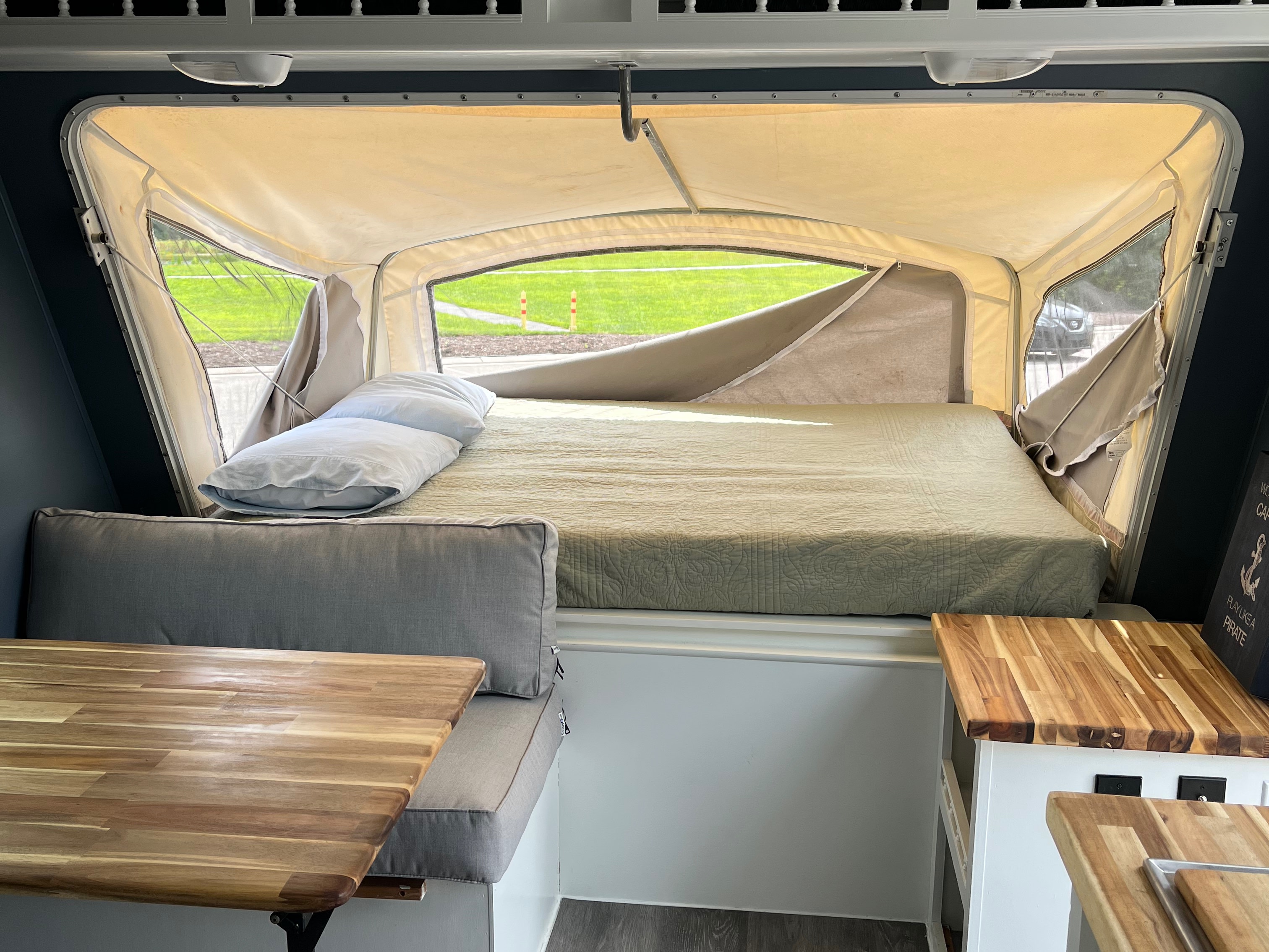 This RV rental features a compact interior: bed with pillows, wood dinette table, and pop-up windows showing green views.. Forest River Surveyor Sport 2013