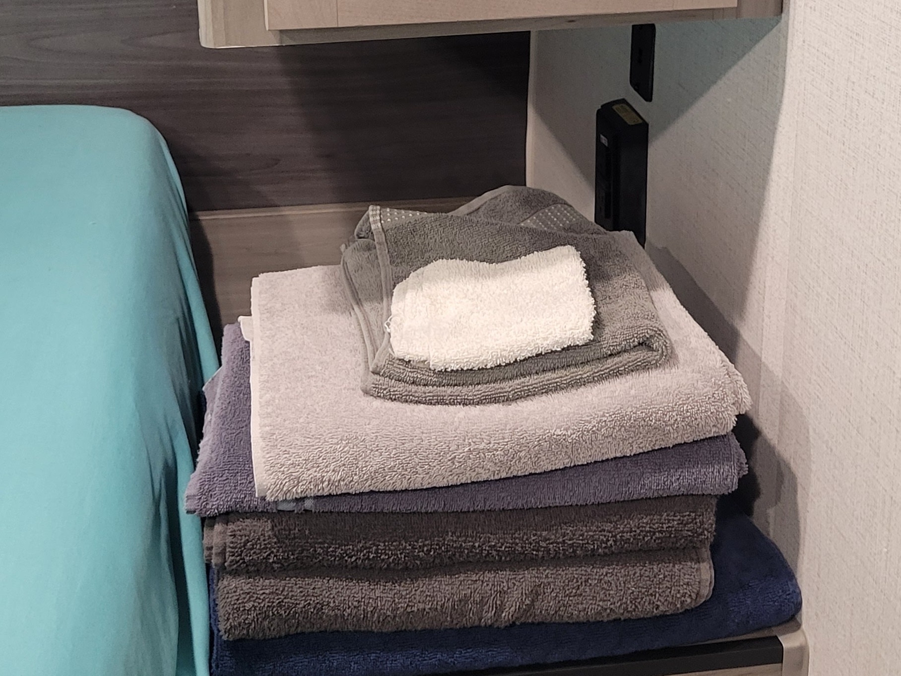 A stack of soft, folded grey and blue towels, neatly arranged on a shelf next to a bed in an RV rental interior.. Keystone RV Hideout 2024