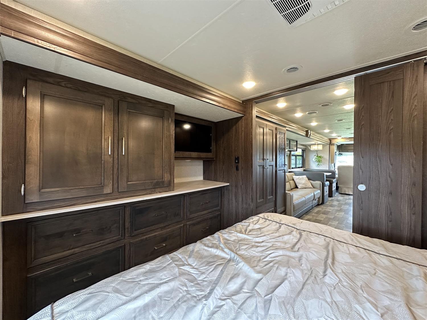 This RV rental features a cozy bedroom with dark wood cabinetry, a TV, and a view into the living area with a sofa and dining.. Coachman Mirada 2018