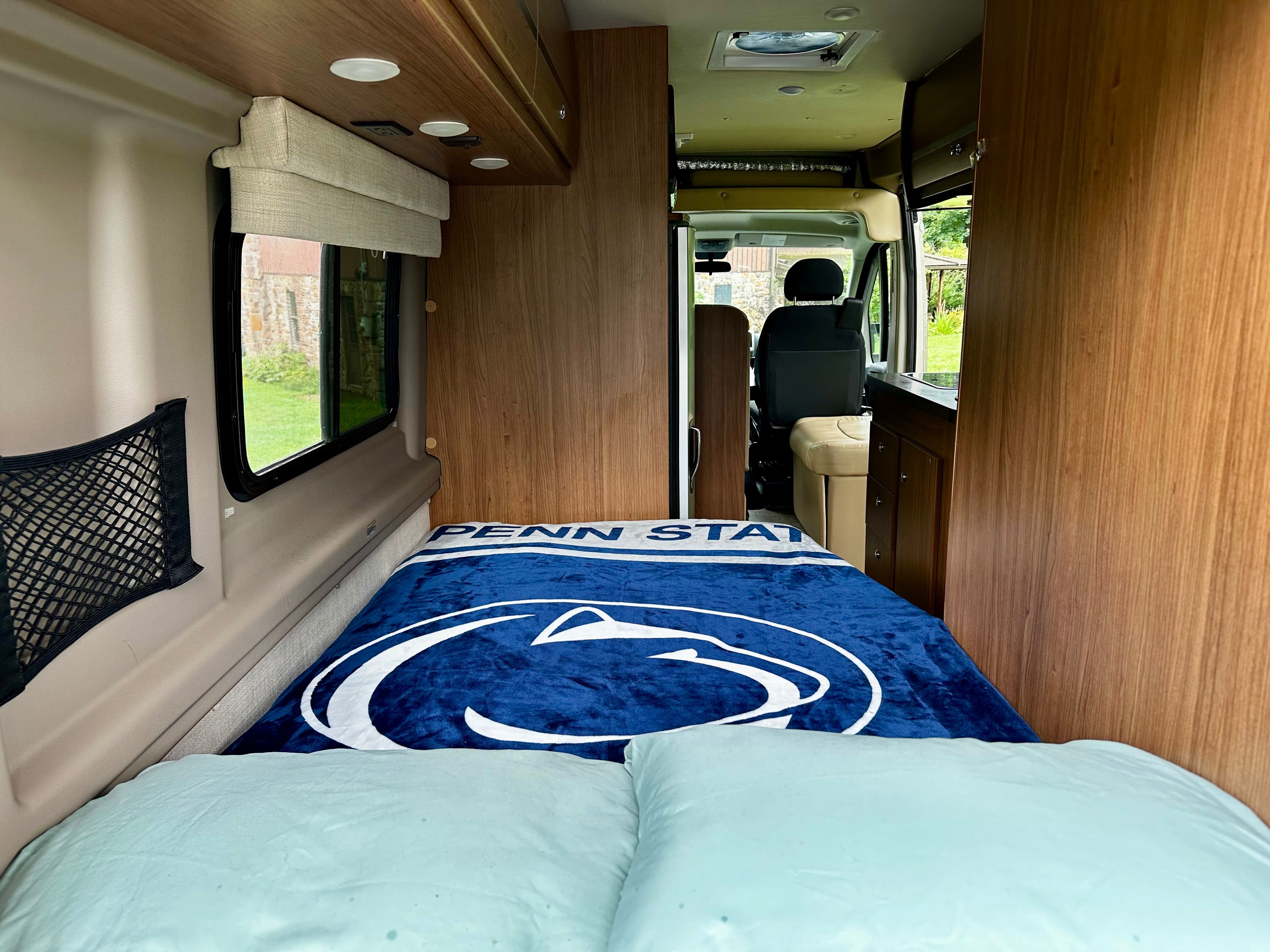 Vibrant Penn State blanket on a cozy bed inside an RV rental, with warm wood interior and window view.. Winnebago Travato 2014