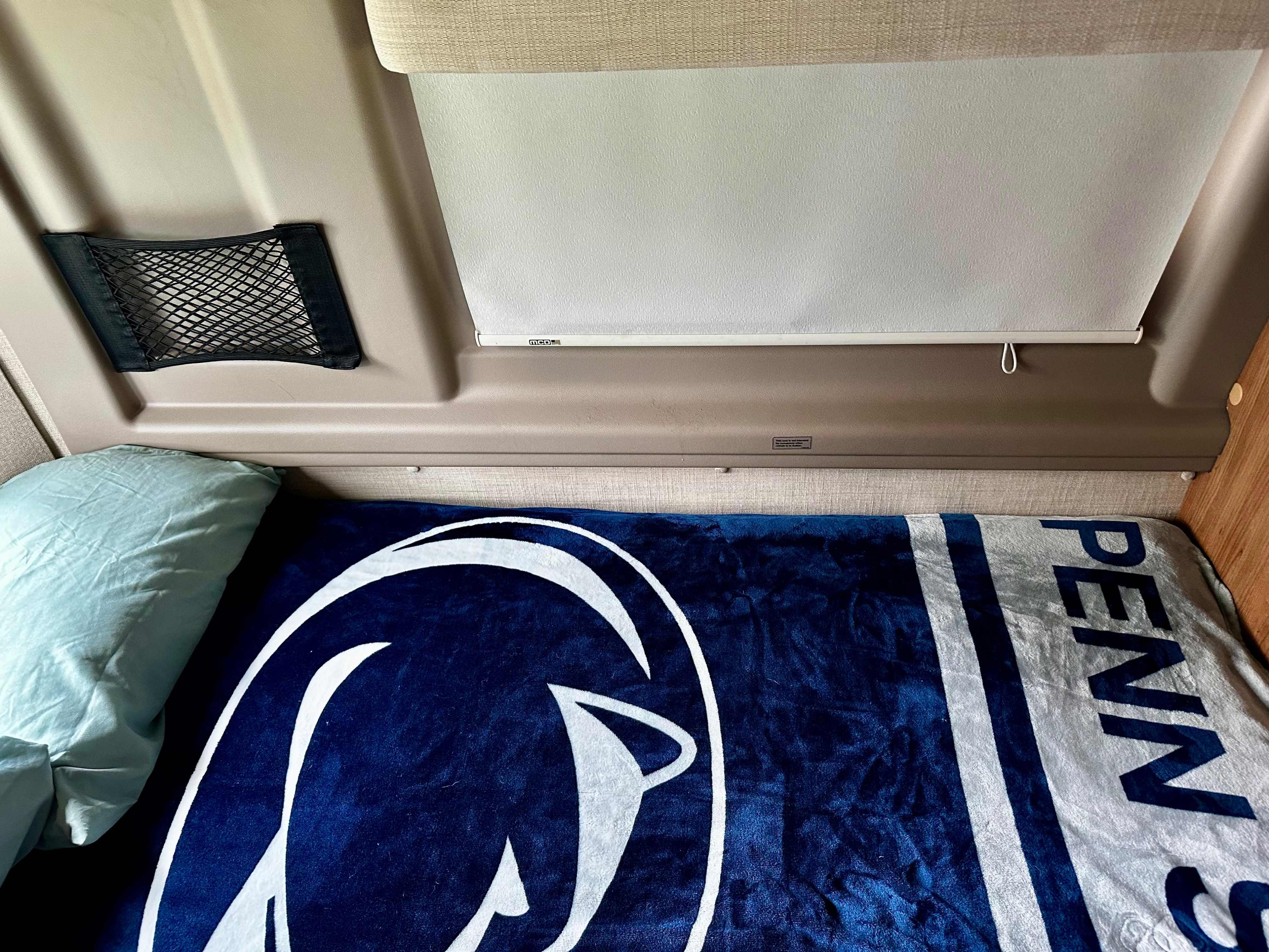 RV rental sleeping nook featuring a Penn State blanket, light blue pillow, mesh storage, and a white window blind.. Winnebago Travato 2014