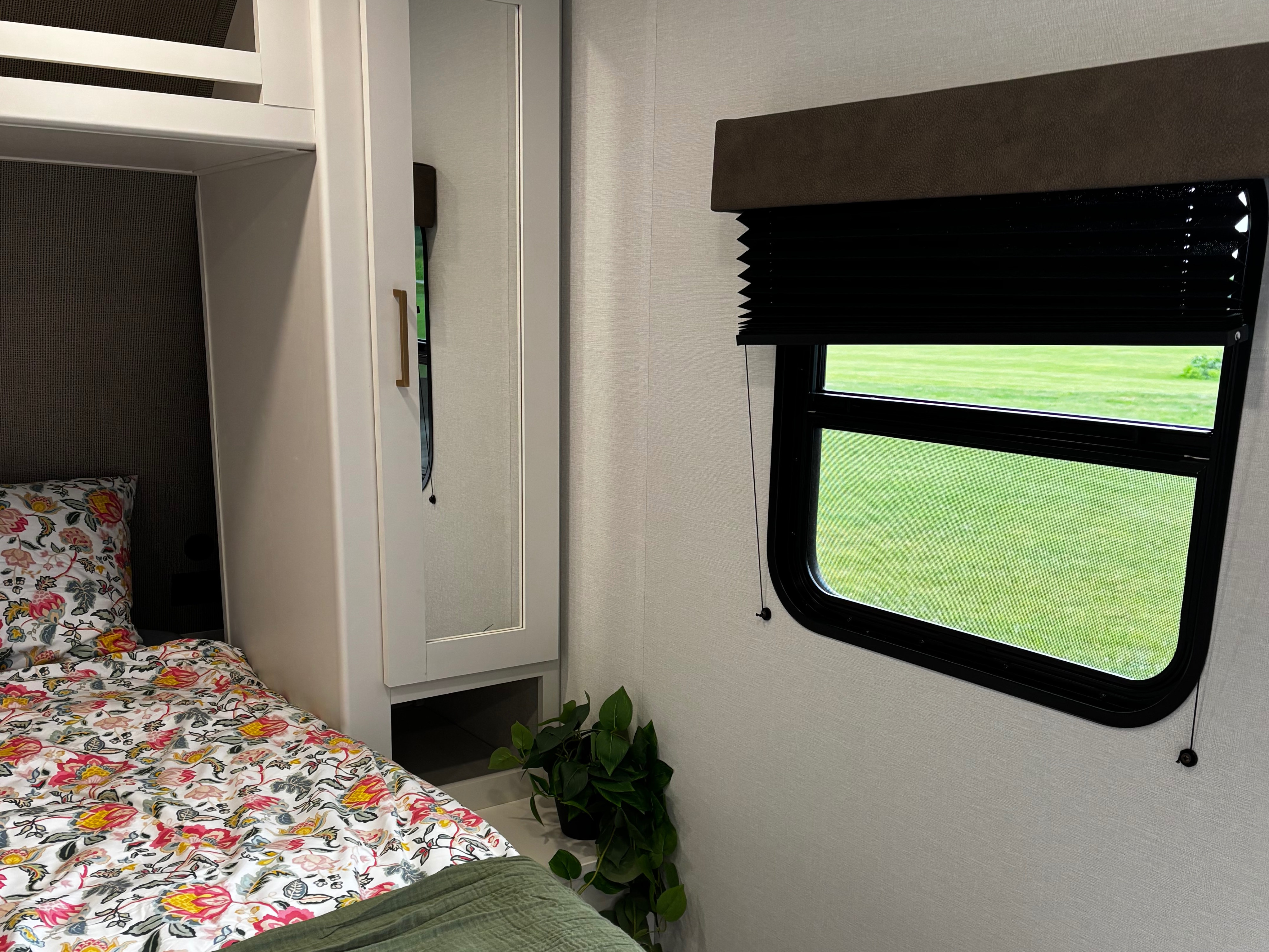 Inside an RV rental: a bunk bed with floral bedding, a mirror, and a window looking out at a vibrant green field.. Grand Design Transcend Xplore 19bhx 2027