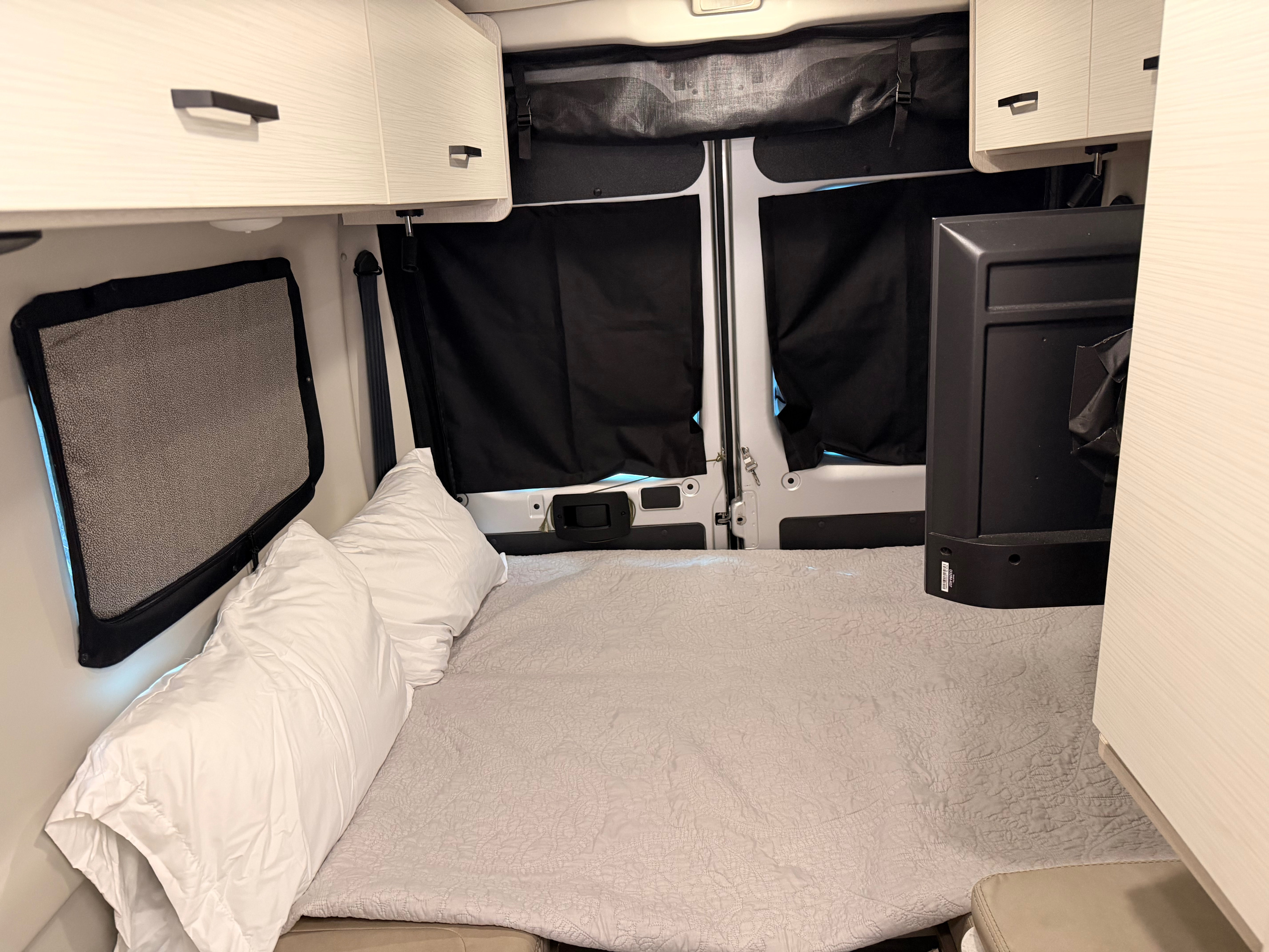 Cozy RV rental interior with a made bed, overhead storage, privacy screens, and TV, ready for comfortable travels.. Dodge Ram 1500 2023