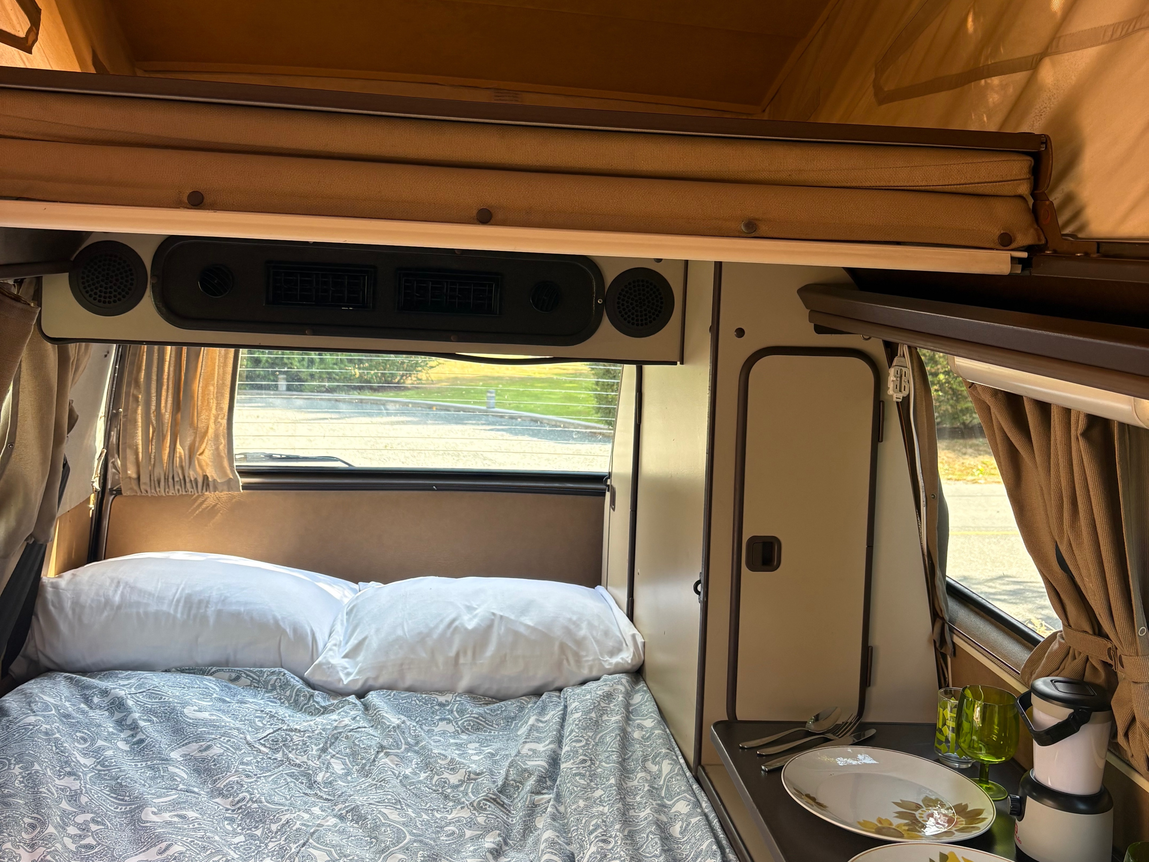 Inside this spacious RV rental: a comfy bed, dining counter with plates, and rear window view. Ideal for travel adventures.. Volkswagen Westfalia 1986