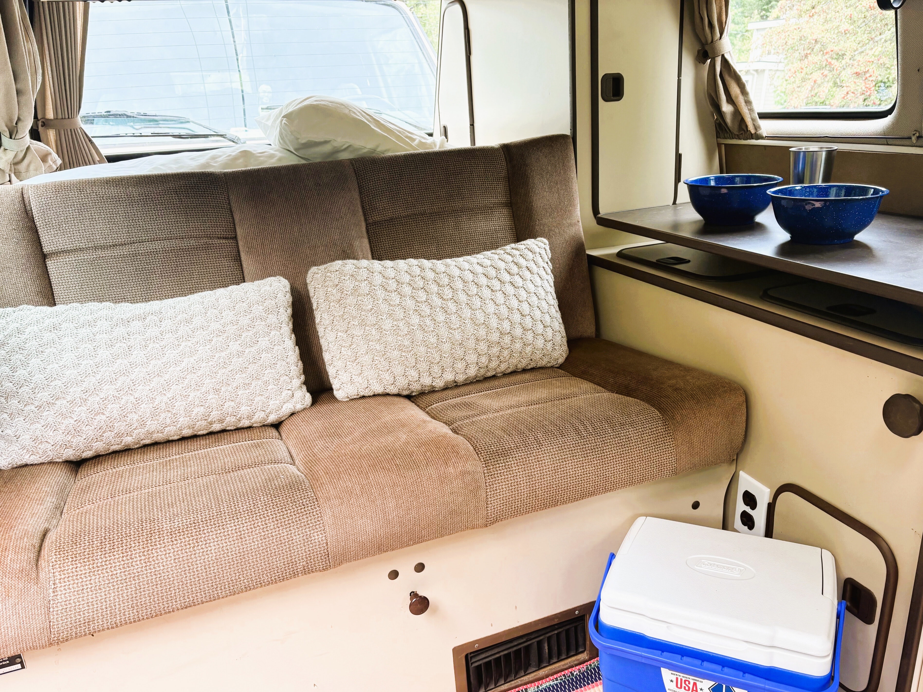 Cozy rv rental interior: A comfortable bench seat, pillows, dishes on a table, and a cooler. Ready for van life adventures!. Volkswagen Westfalia 1985