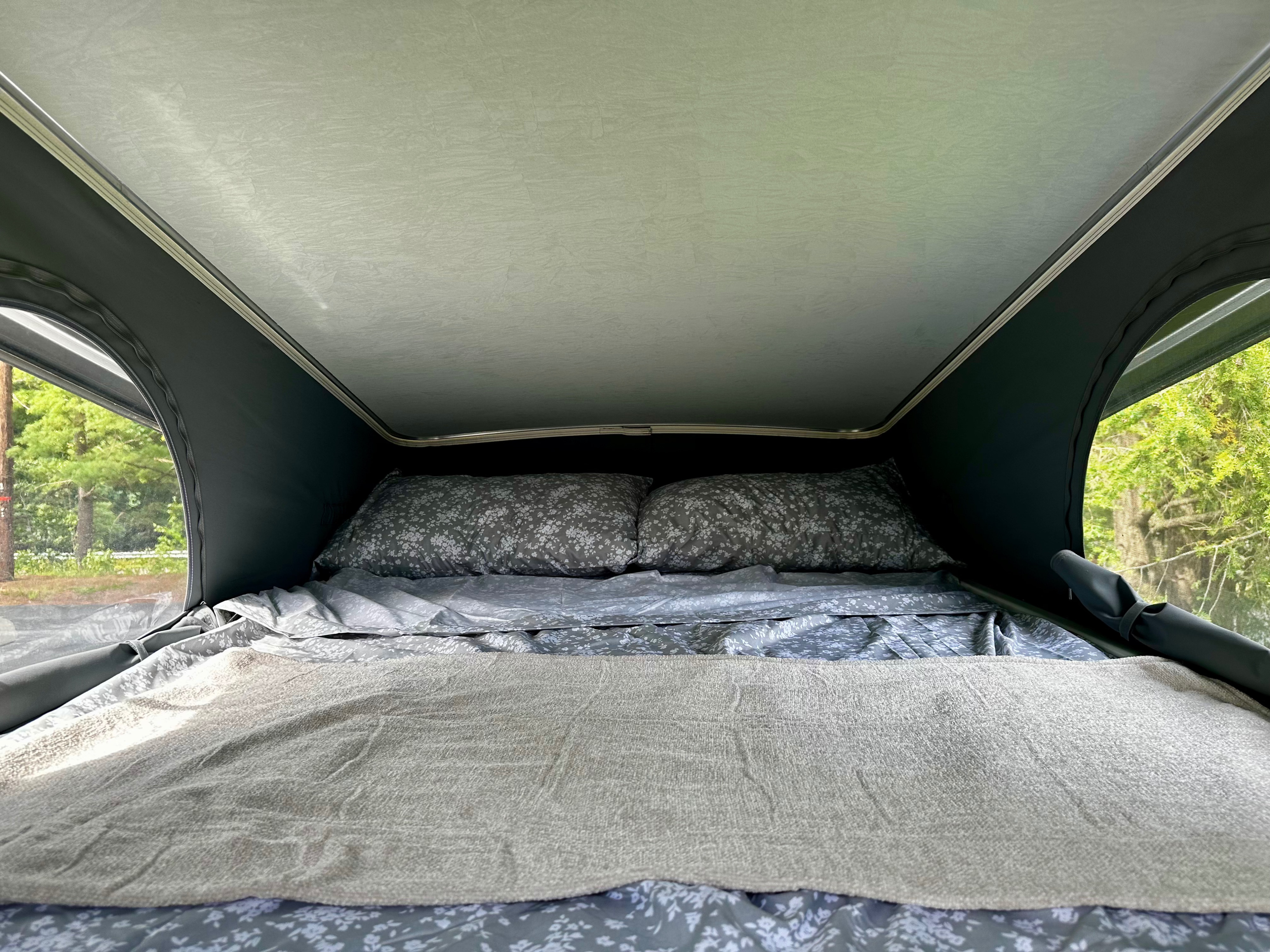 Inside an RV rental camper van: a cozy sleeping area with bed, pillows, and blankets. Pop-top windows show trees.. Winnebago Solis 59PX 2023