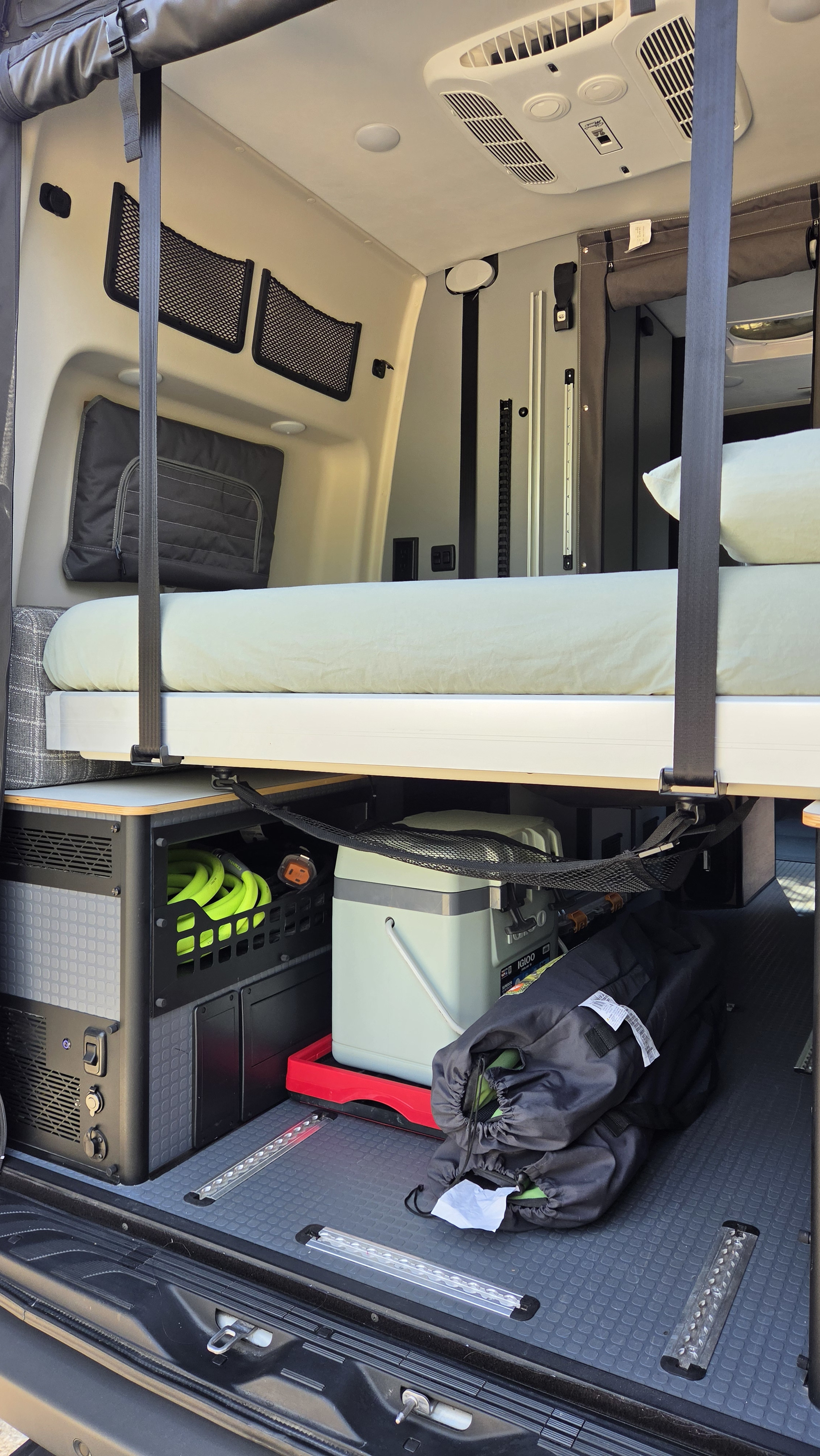 RV rental interior showing an elevated bed above organized storage, a cooler, and gear on a textured floor, with overhead AC.. Winnebago Revel 2026