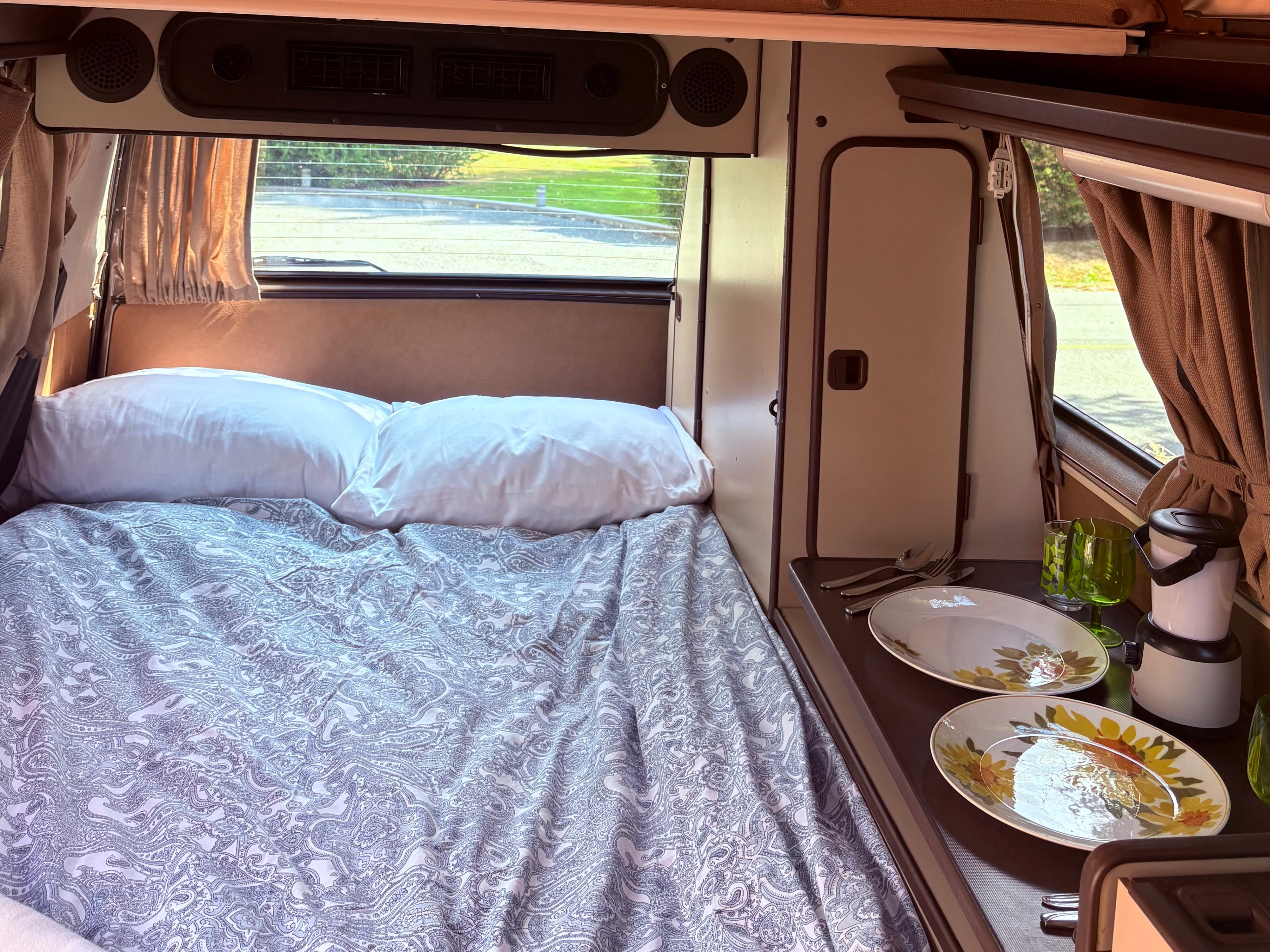 Cozy rv rental camper van interior with a comfy bed, patterned bedding, and a small dining setup with plates and glasses.. Volkswagen Westfalia 1986