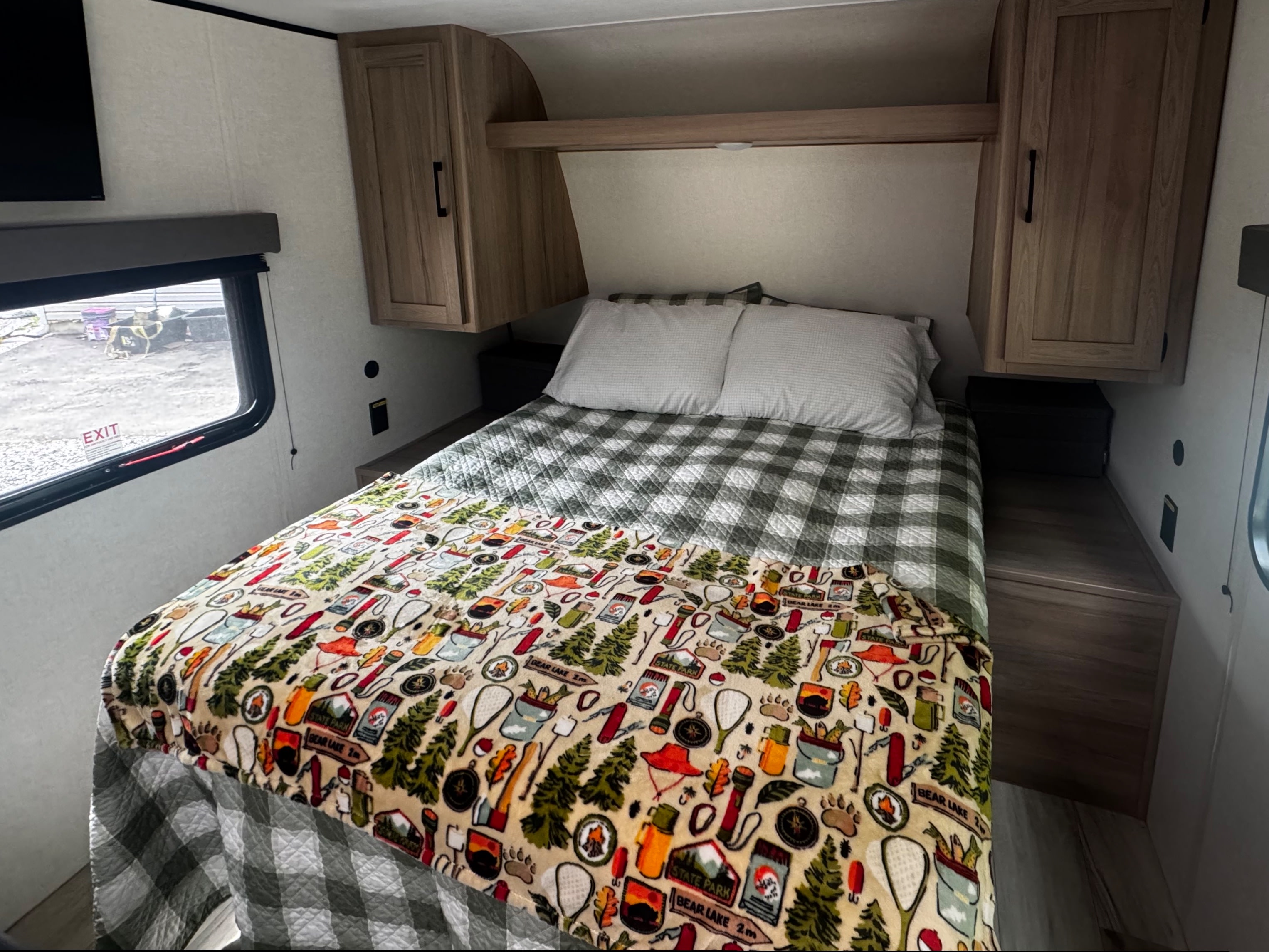 Cozy RV rental bedroom with a bed, plaid quilt, and fun camping-themed blanket, featuring overhead storage and a window.. Keystone RV Coleman 2025