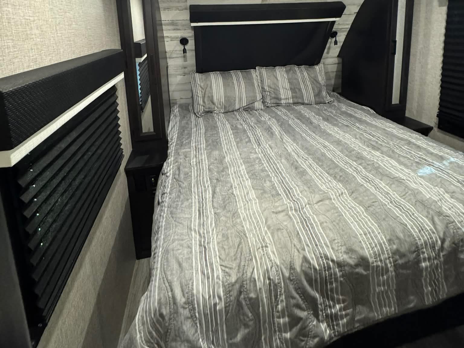 Modern RV rental bedroom with striped bed, dark headboard, and window blinds. Ideal for travel comfort.. Jayco White Hawk 2022