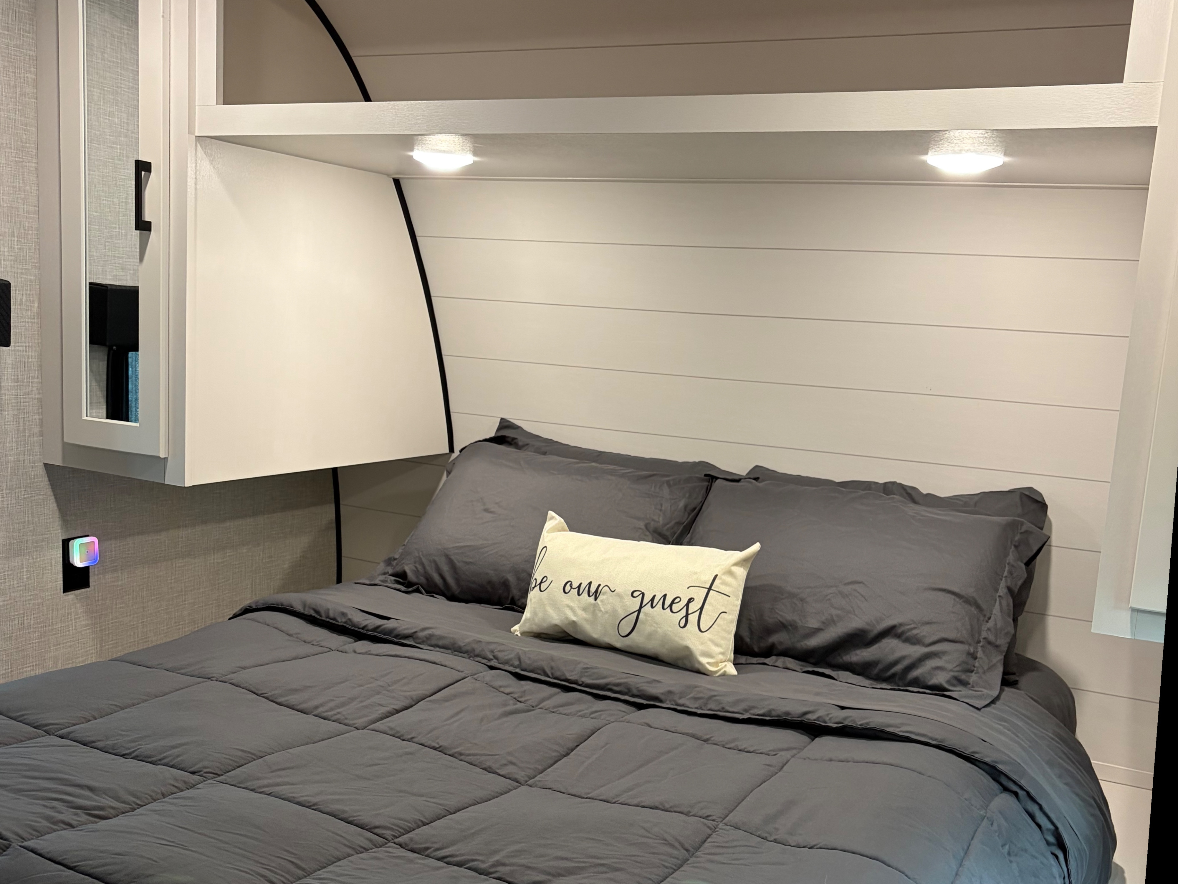 This RV rental offers a cozy bedroom with a dark gray bed, decorative pillow, shiplap-style headboard, overhead storage, and ambient lighting.. Jayco Jay Flight SLX 2024
