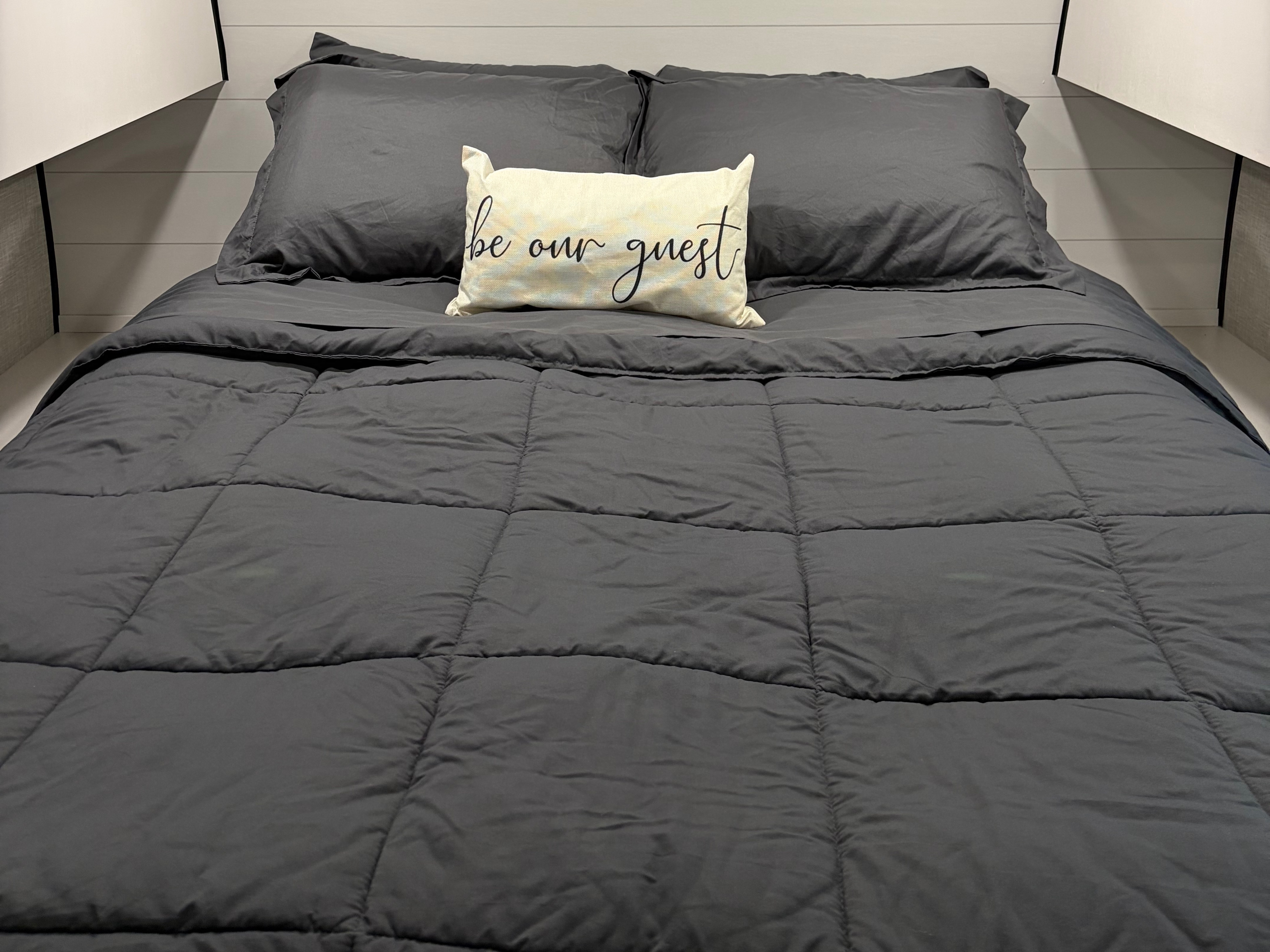 An inviting, neatly made bed in an RV rental, featuring dark gray bedding and a "be our guest" pillow.. Jayco Jay Flight SLX 2024