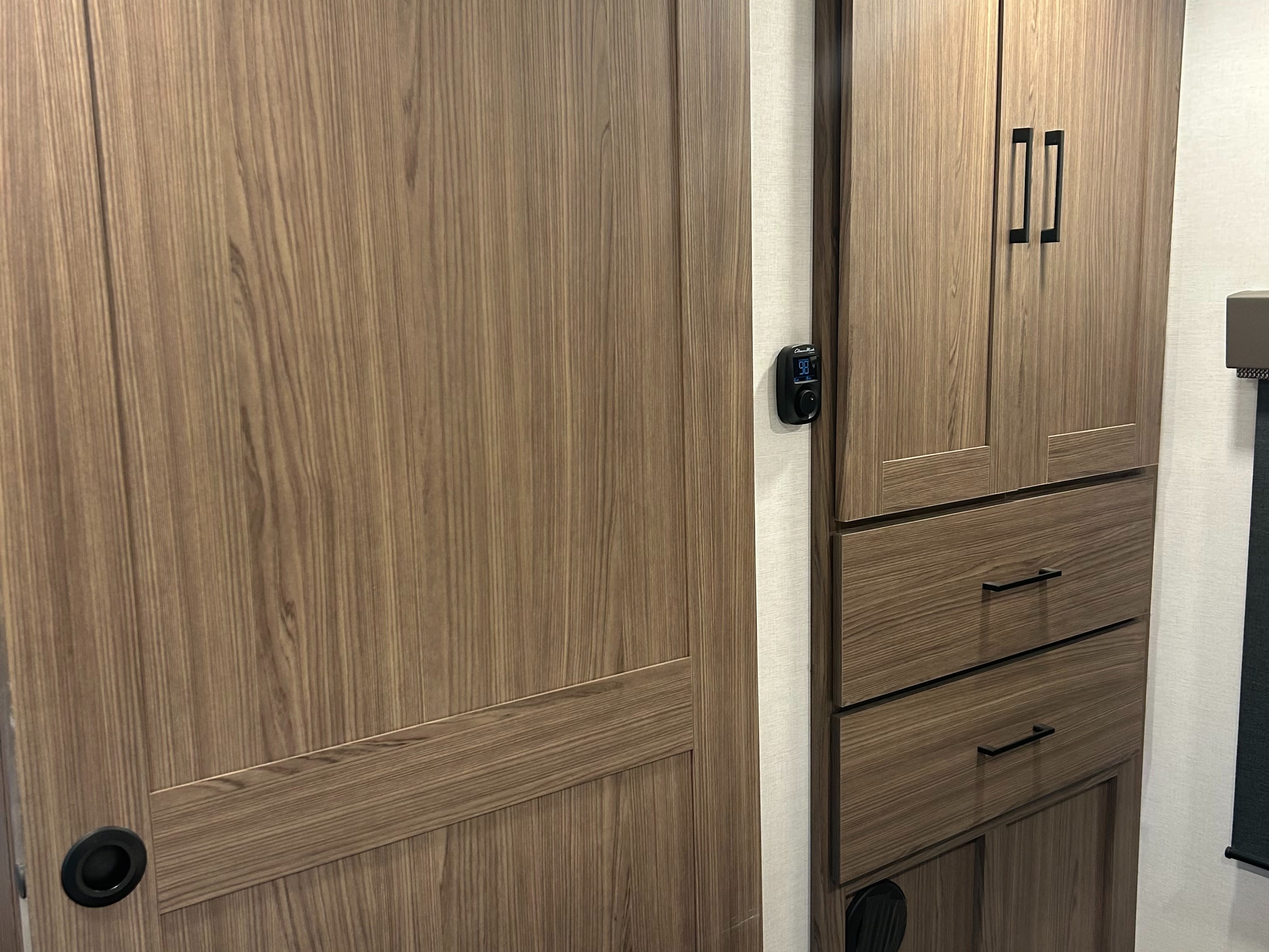 Modern RV rental interior: wood-grain sliding door, digital thermostat (98F), and storage cabinet with drawers.. Alliance RV Delta 2025