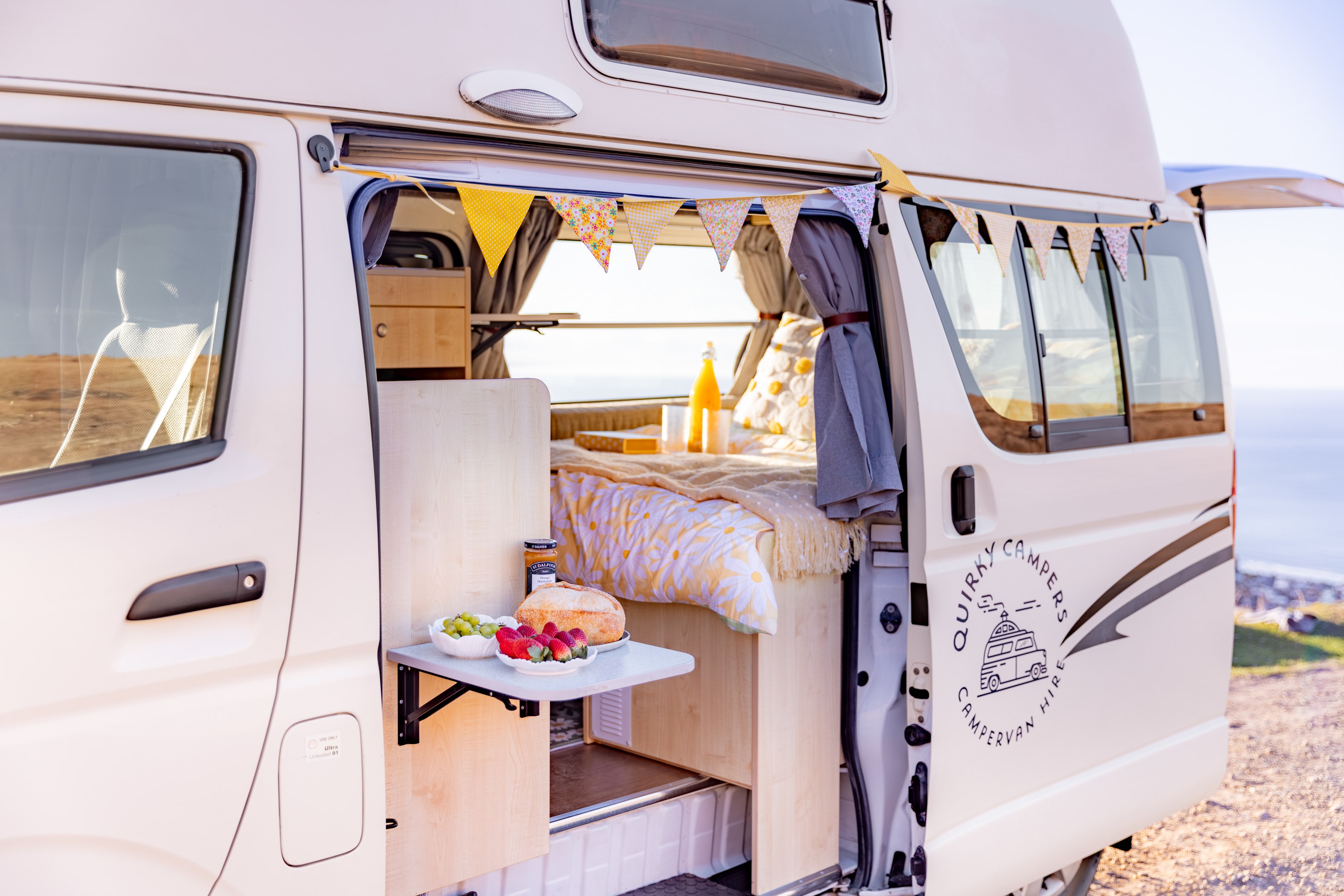 Quirky Campers RV rental: A cozy campervan's interior and picnic table, open to a scenic ocean view, perfect for adventure.. Toyota Hiace 2013