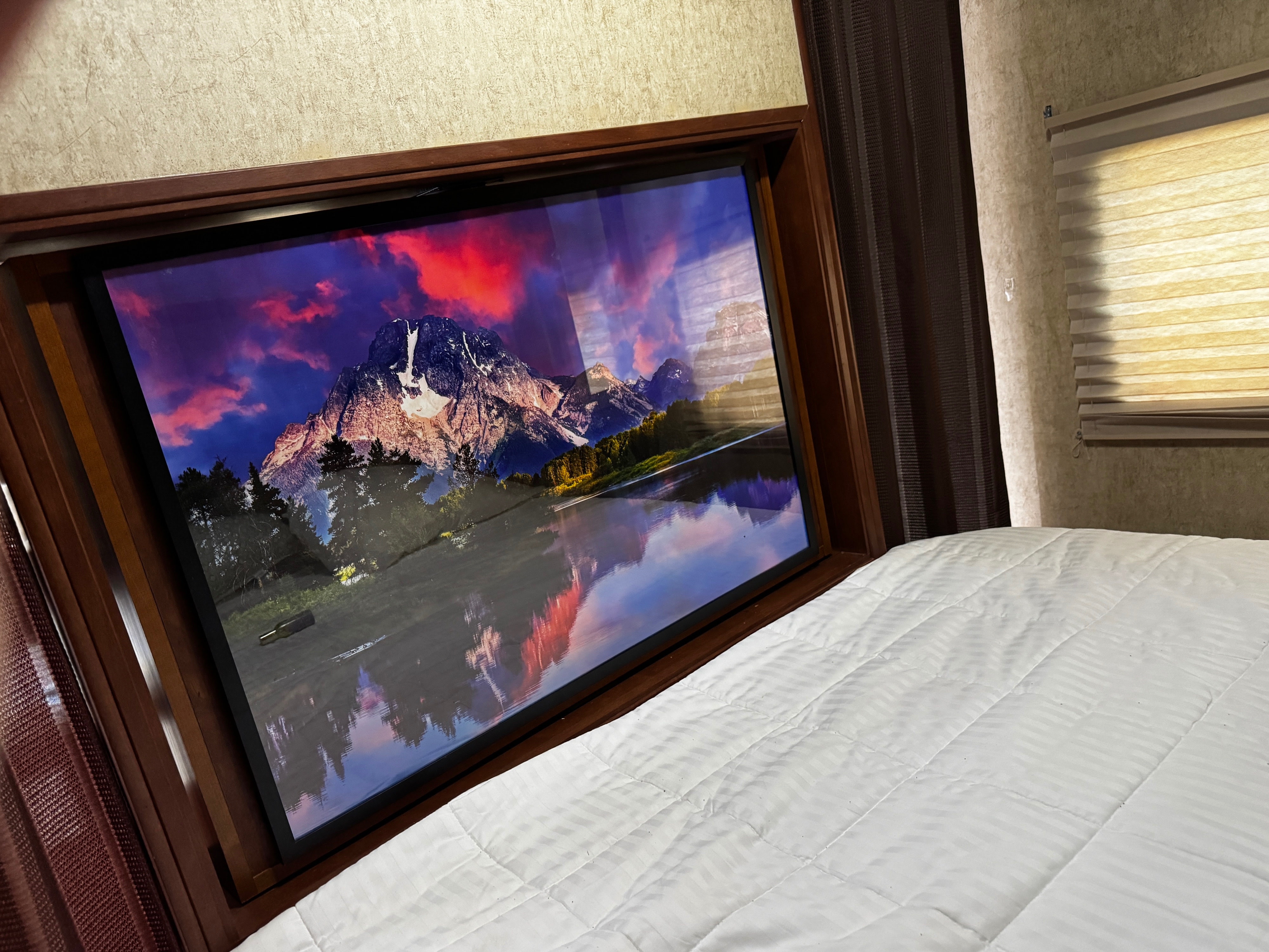 RV rental bedroom: Relax on a comfortable white bed facing a large TV displaying a vibrant mountain landscape with a reflective lake.. Heartland RVs Mallard 2014