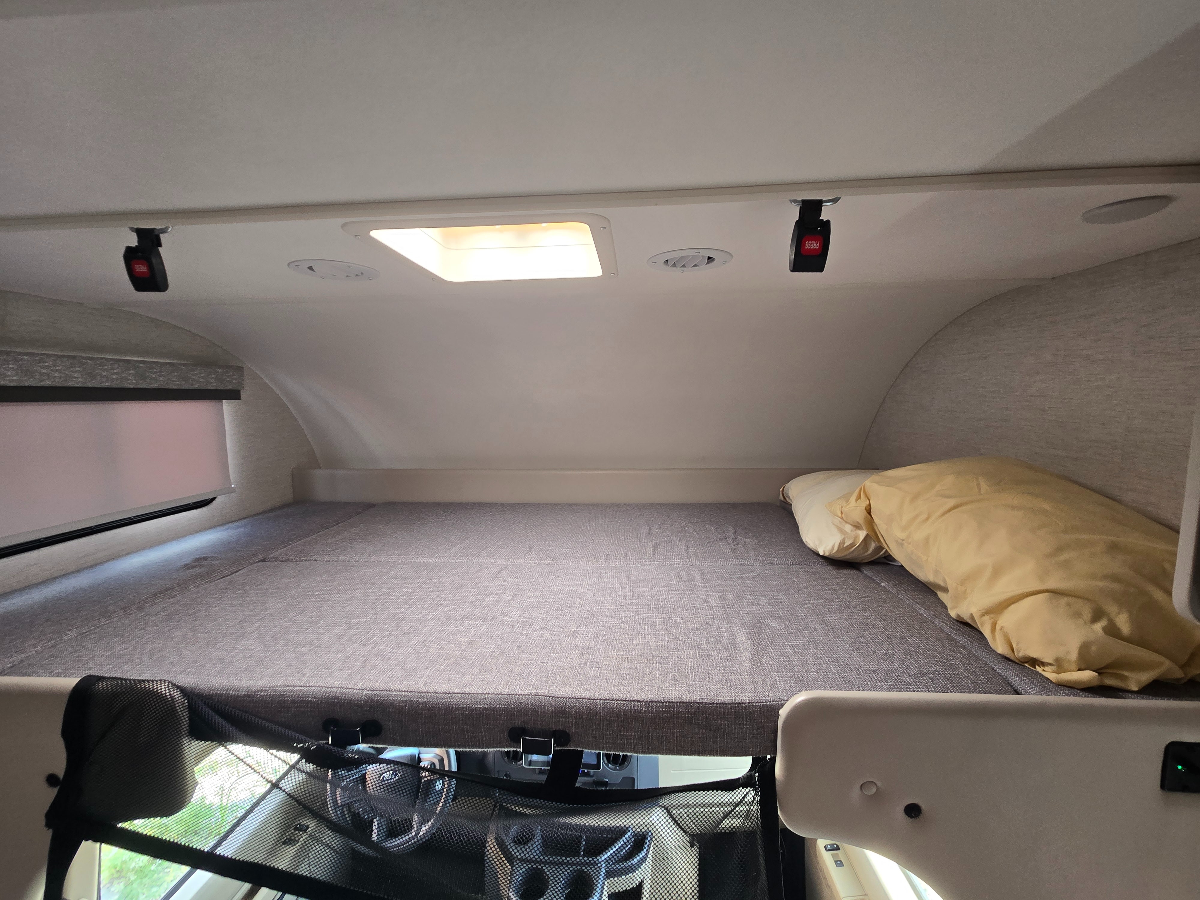 Cozy overhead bunk in an RV rental, featuring a mattress, pillows, a window, and a safety net above the driver's cabin.. Thor Four Winds 2025
