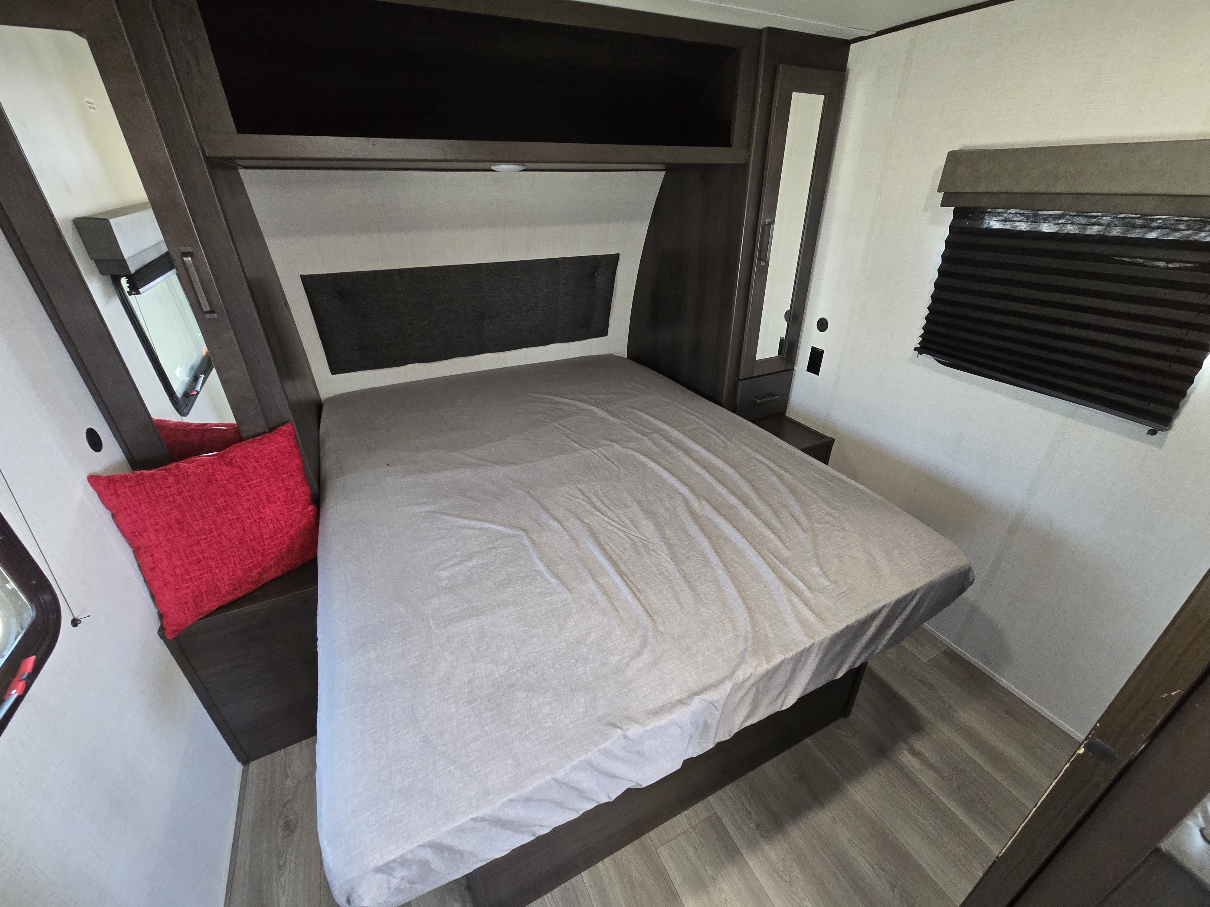 RV rental bedroom: A bed with light gray sheets, dark headboard, red pillow, built-in dark wood storage, and window with blind.. Grand Design Transcend Xplor 2023