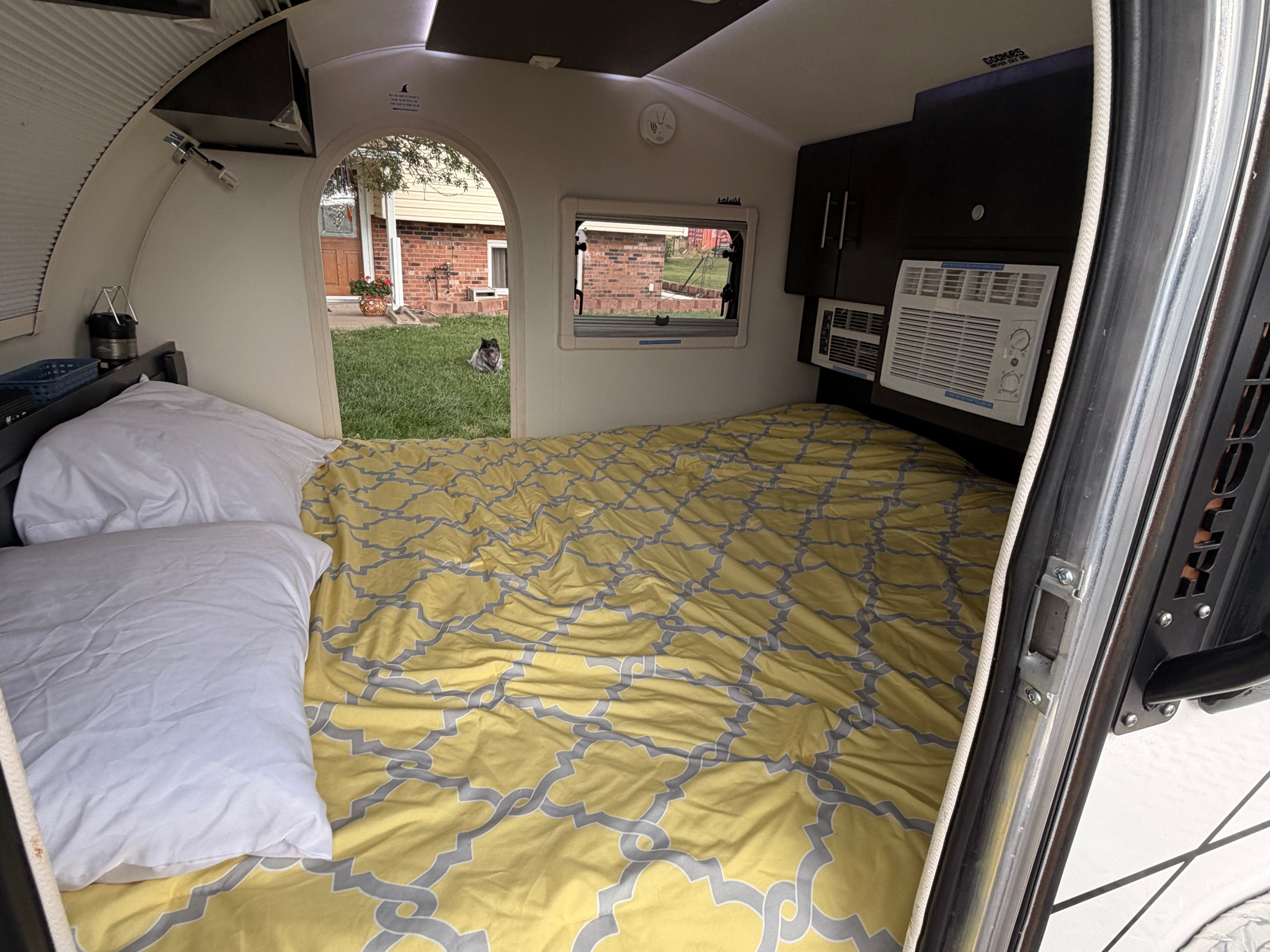 Inside a cozy teardrop camper rv rental: bed with patterned comforter, AC unit, and a dog watching from the yard.. nuCamp T@G 2021