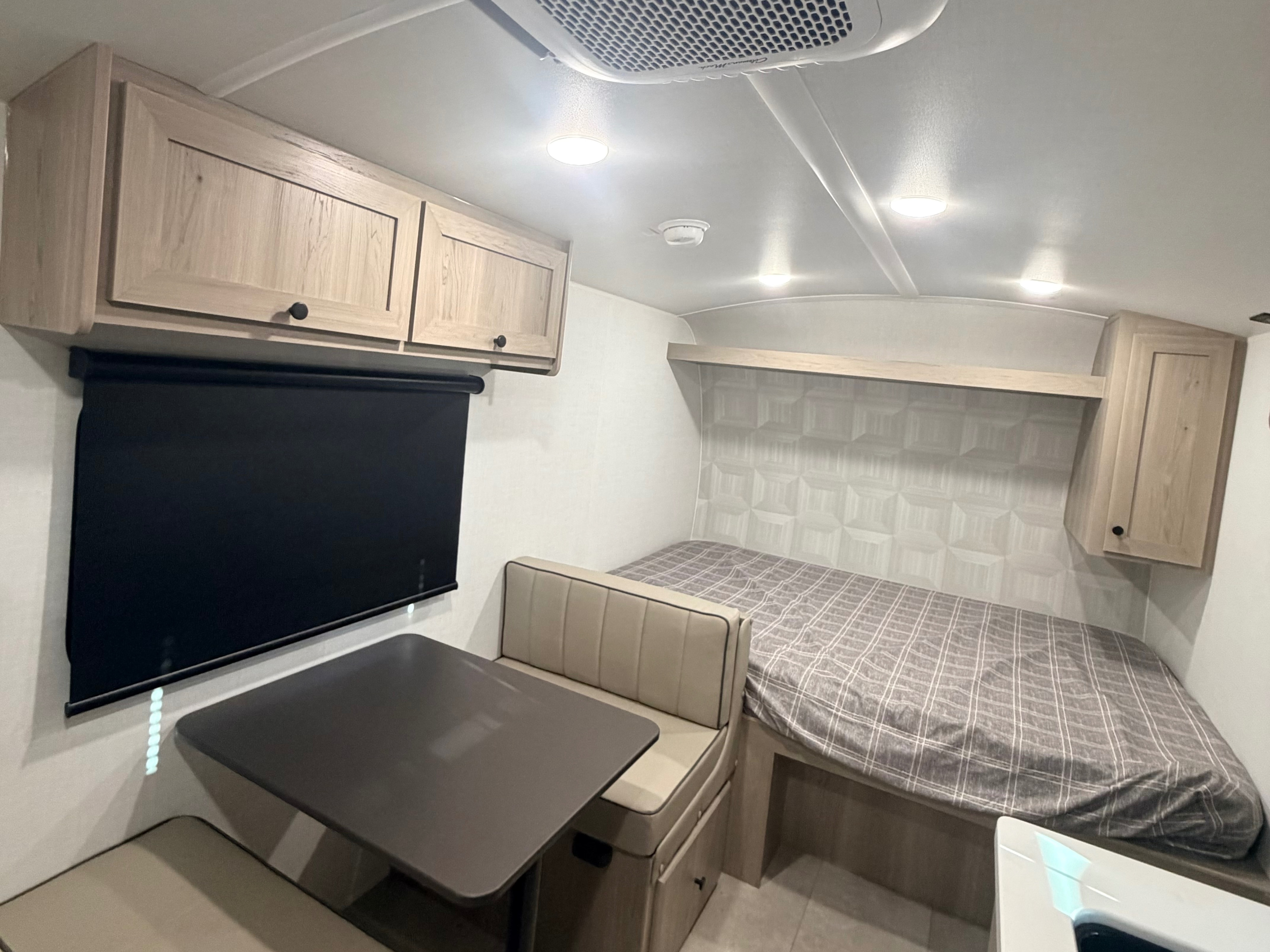 Compact RV rental interior featuring a dinette with bench seating, black roller blind, and bed with plaid bedding. Overhead cabinets provide storage in this efficient space.. Forest River Rockwood Geo Pro 17LE 2026
