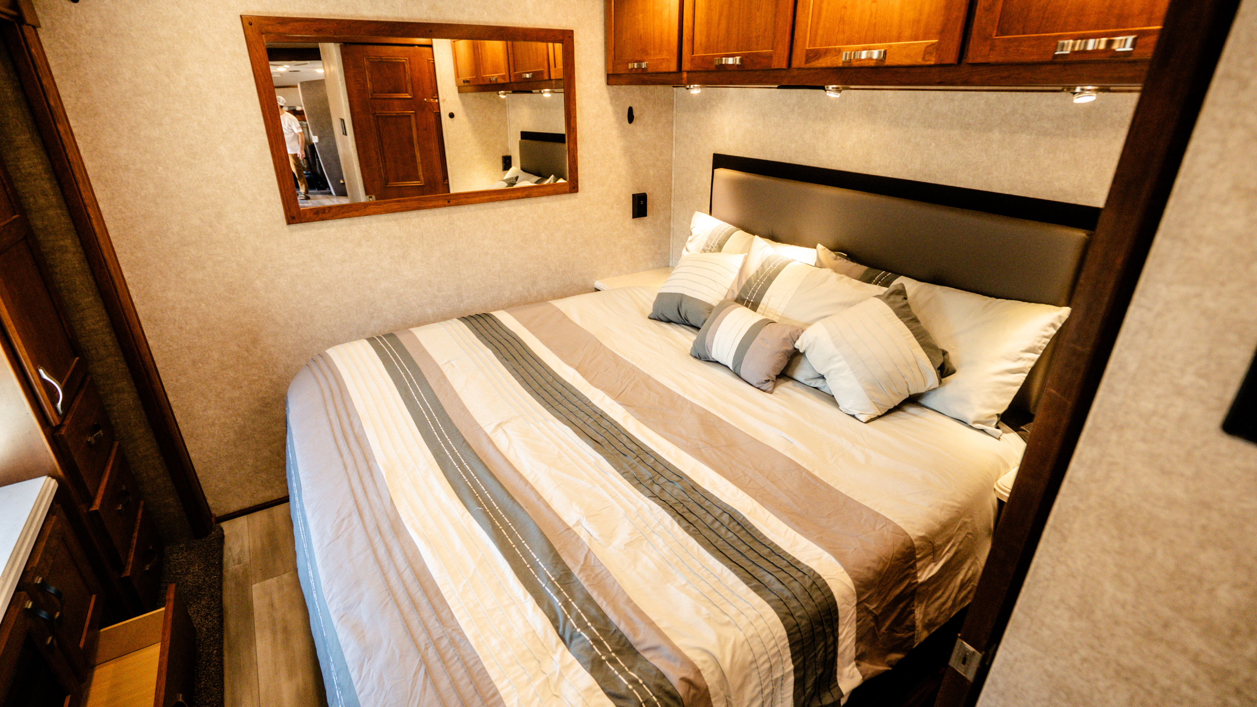 This cozy RV rental bedroom features a comfy bed with striped bedding, stylish wooden cabinets, and a convenient mirror.. Renegade Custom Coaches Valencia 2022