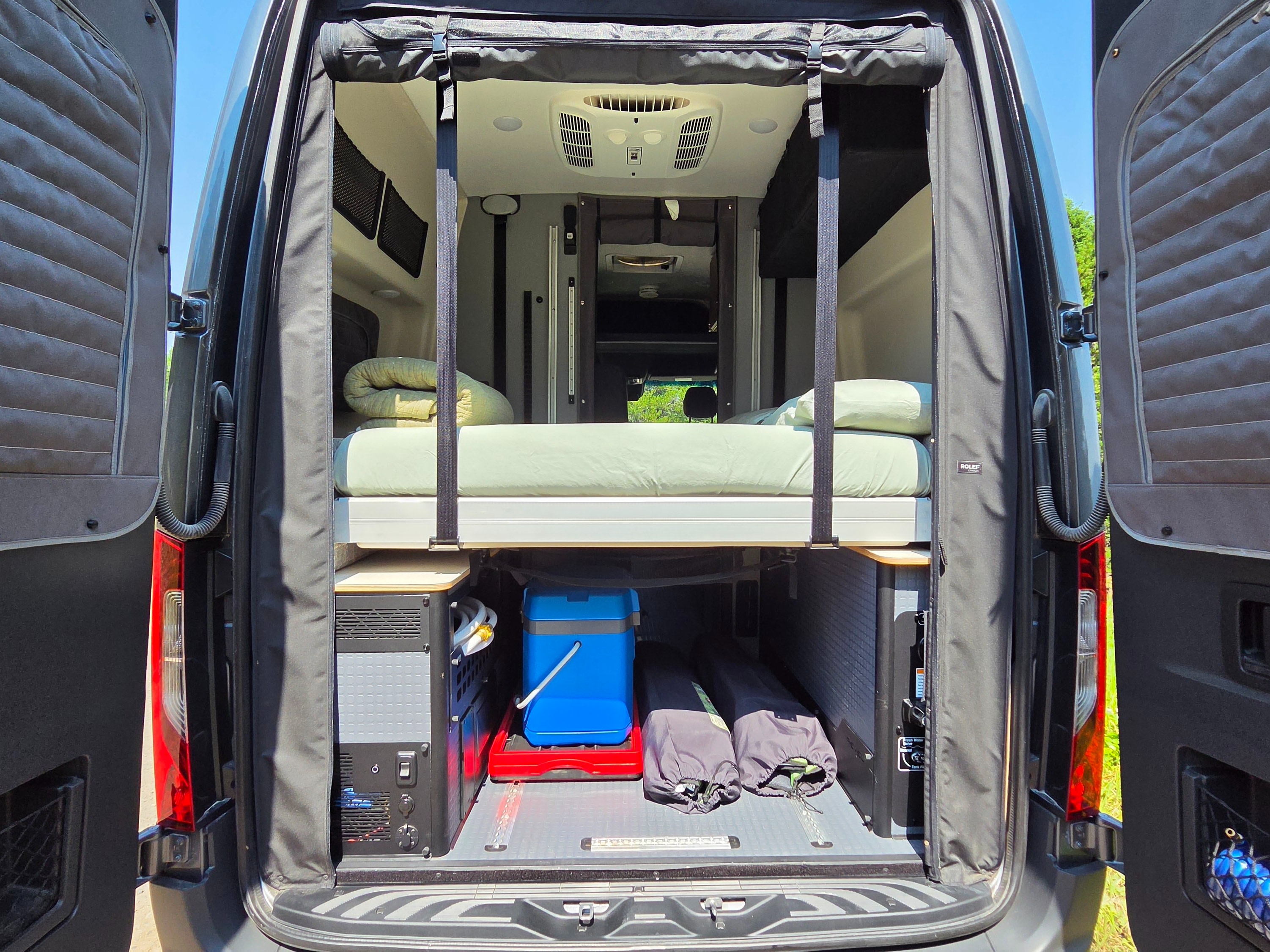 RV rental: Open rear doors of a camper van show a cozy elevated bed, with organized storage below, including a cooler and gear.. Winnebago Revel 2025