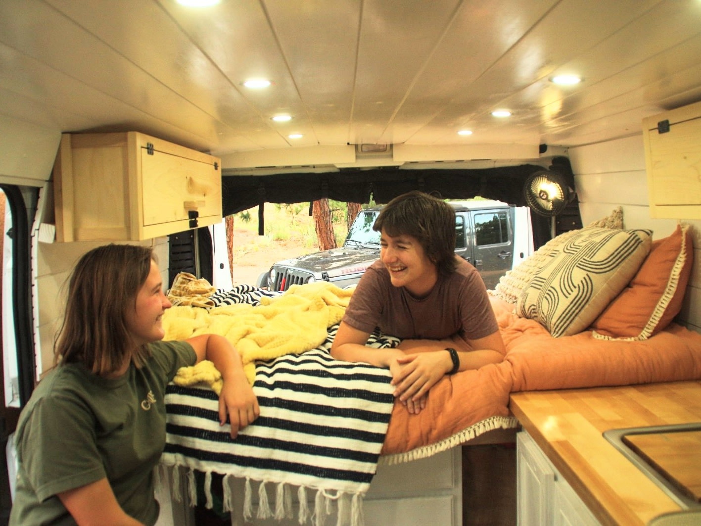 Two young adults smile inside a cozy rv rental camper van, featuring a bed, kitchen counter, and a Jeep visible outside.. Ram Promaster 1500 2017