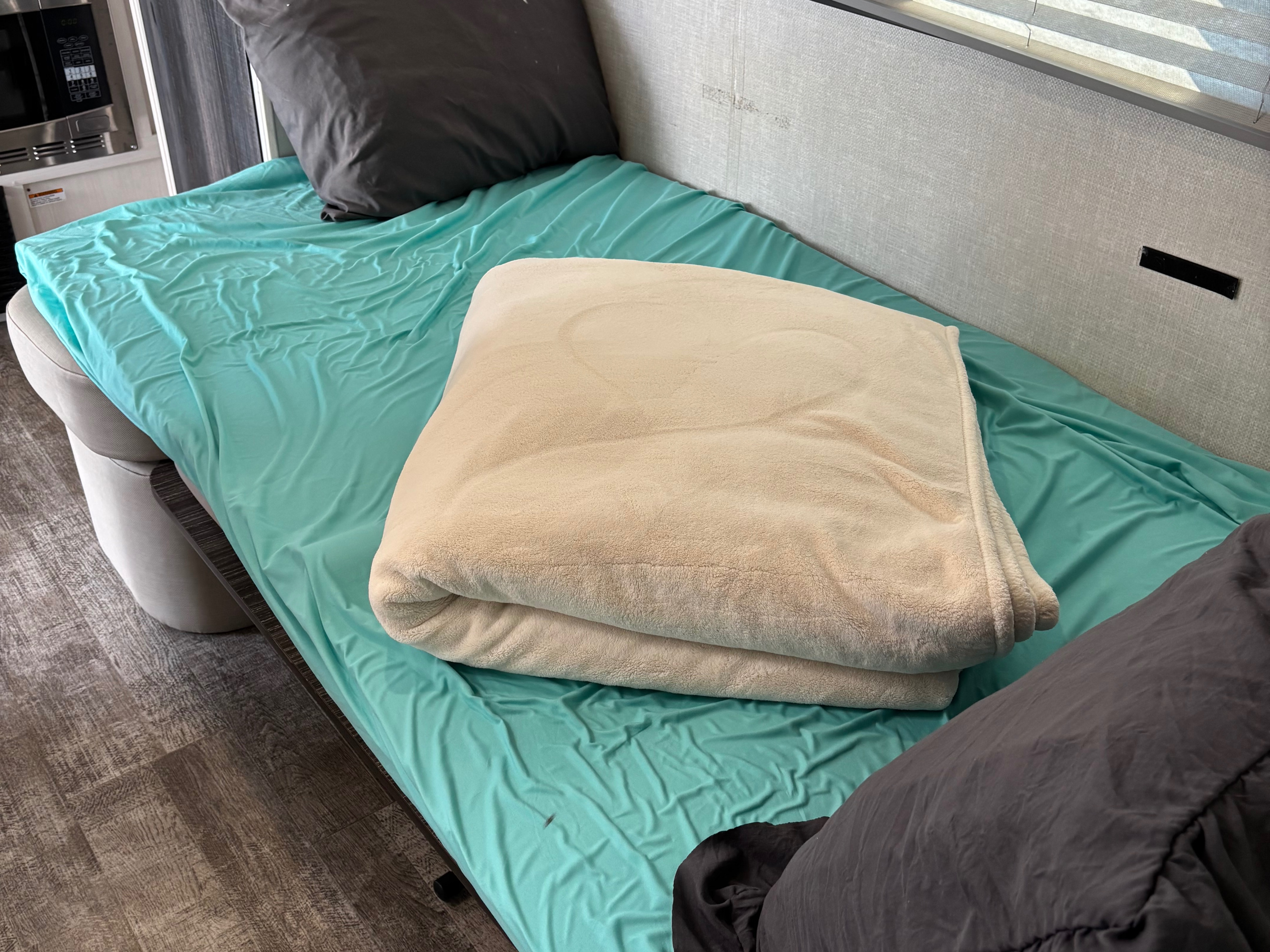 Cozy bed setup in an RV rental, featuring a teal sheet, grey pillow, and cream blanket. A microwave is visible in the background.. Forest River 195 RPod 2021