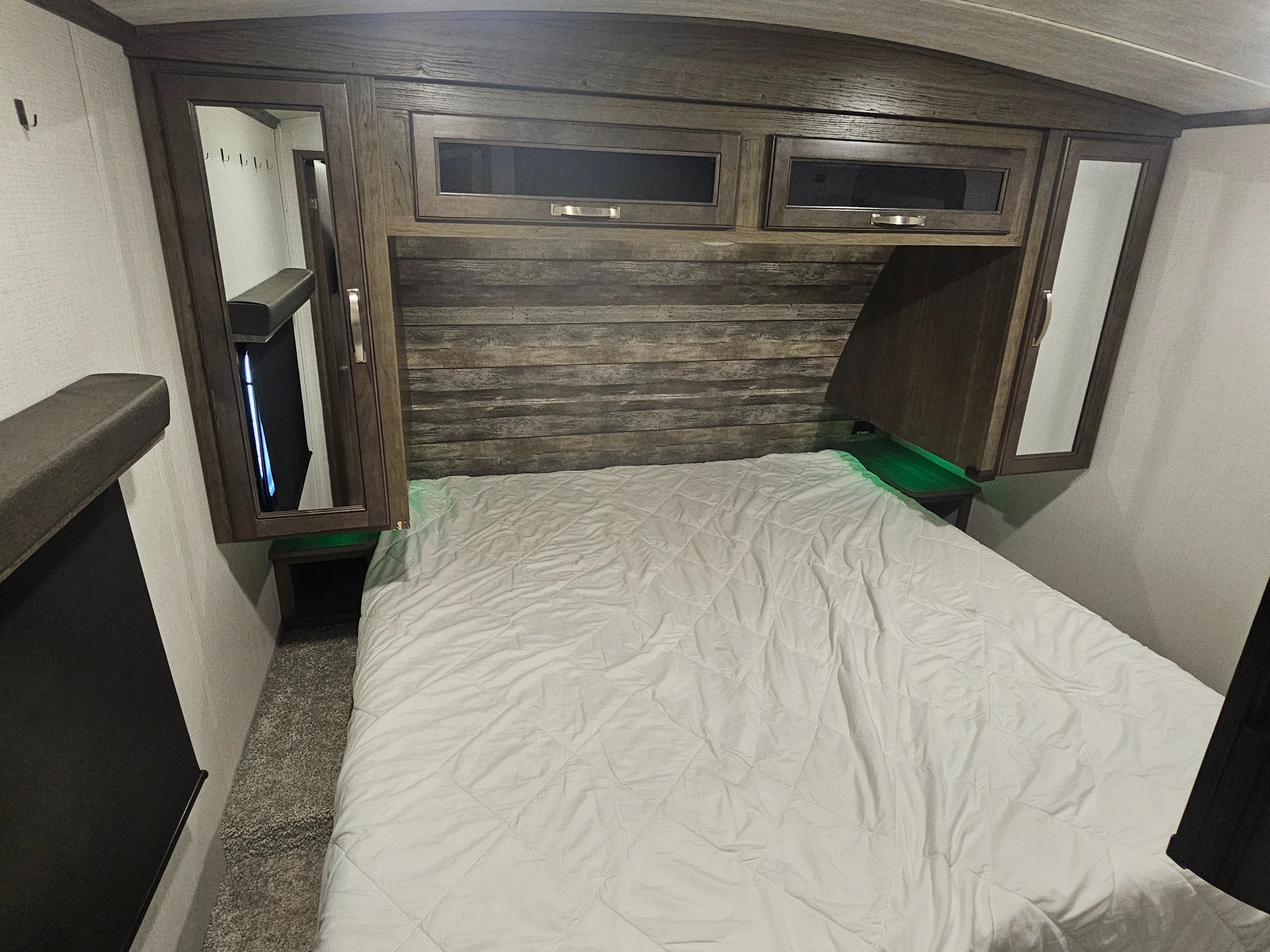 RV rental bedroom featuring a comfy bed with white comforter, wooden headboard, overhead cabinets, and side wardrobe.. Keystone RV Cougar 2022