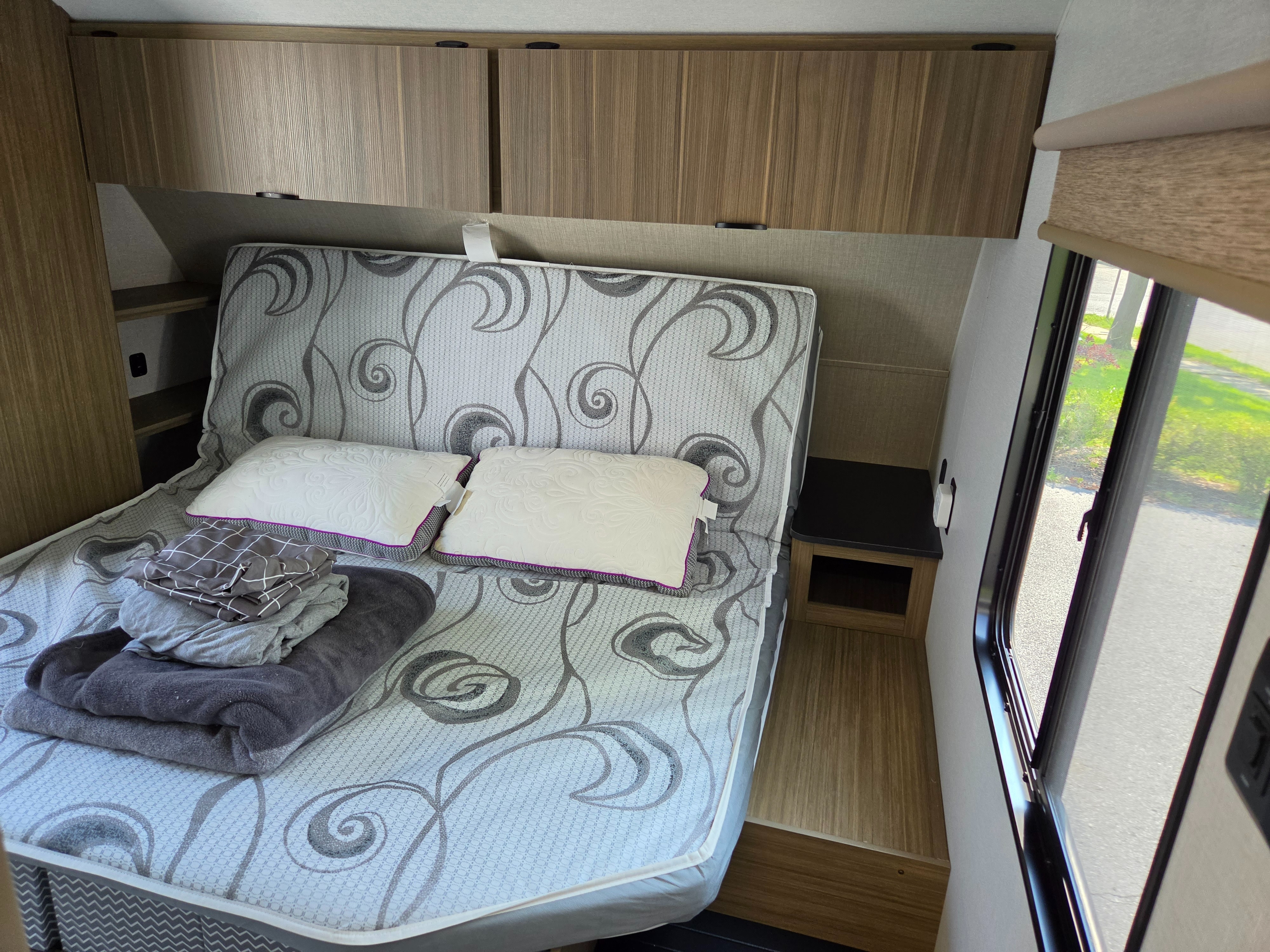 RV rental bedroom showing a bed with a patterned mattress, pillows, folded blankets, overhead cabinets, and a window.. Forest River Wildwood X-Lite 2025