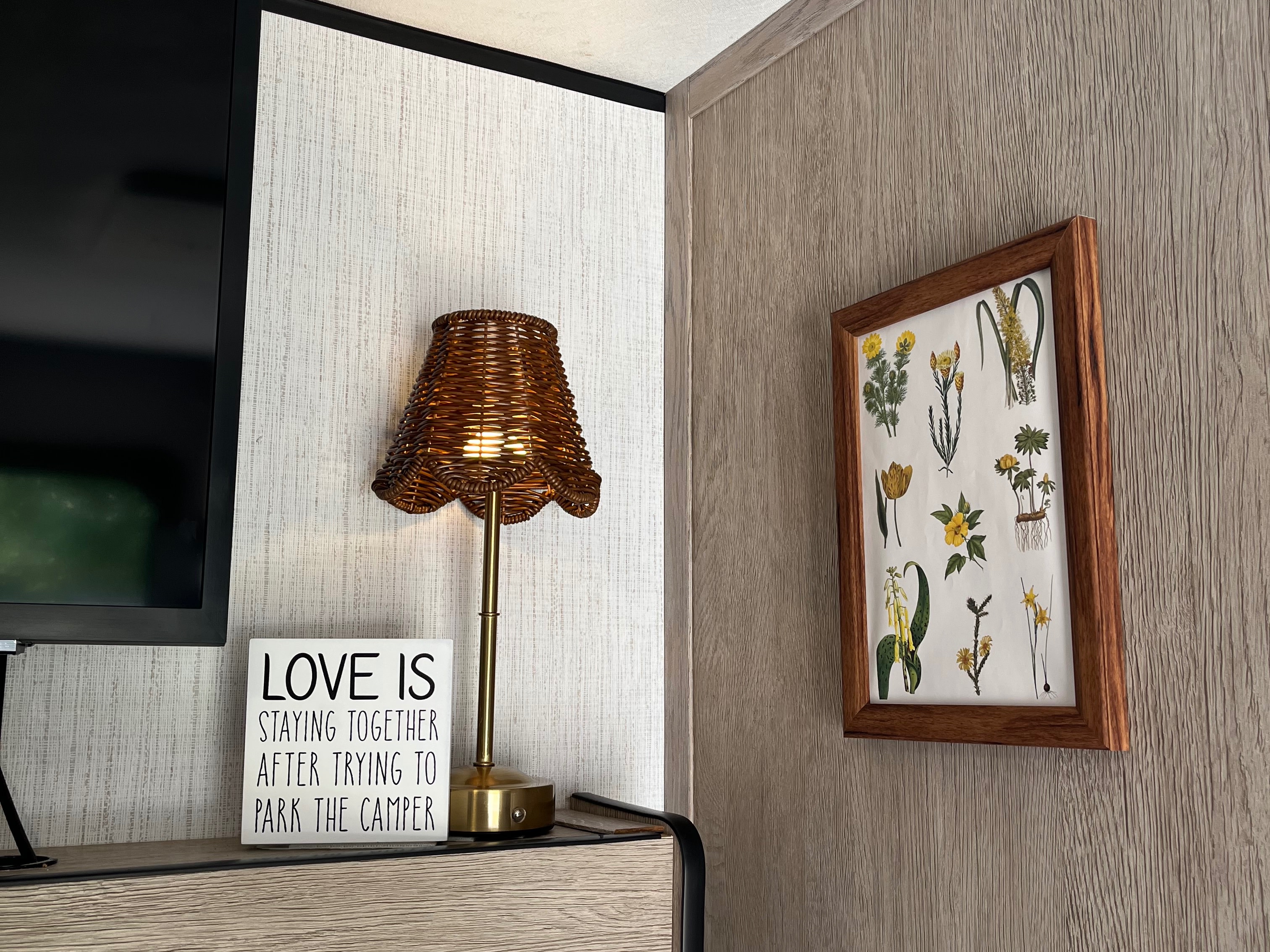 Cozy RV rental interior with TV, woven lamp, "Love is..." sign about parking a camper, & botanical art on textured walls.. Heartland RVs Prowler 2021