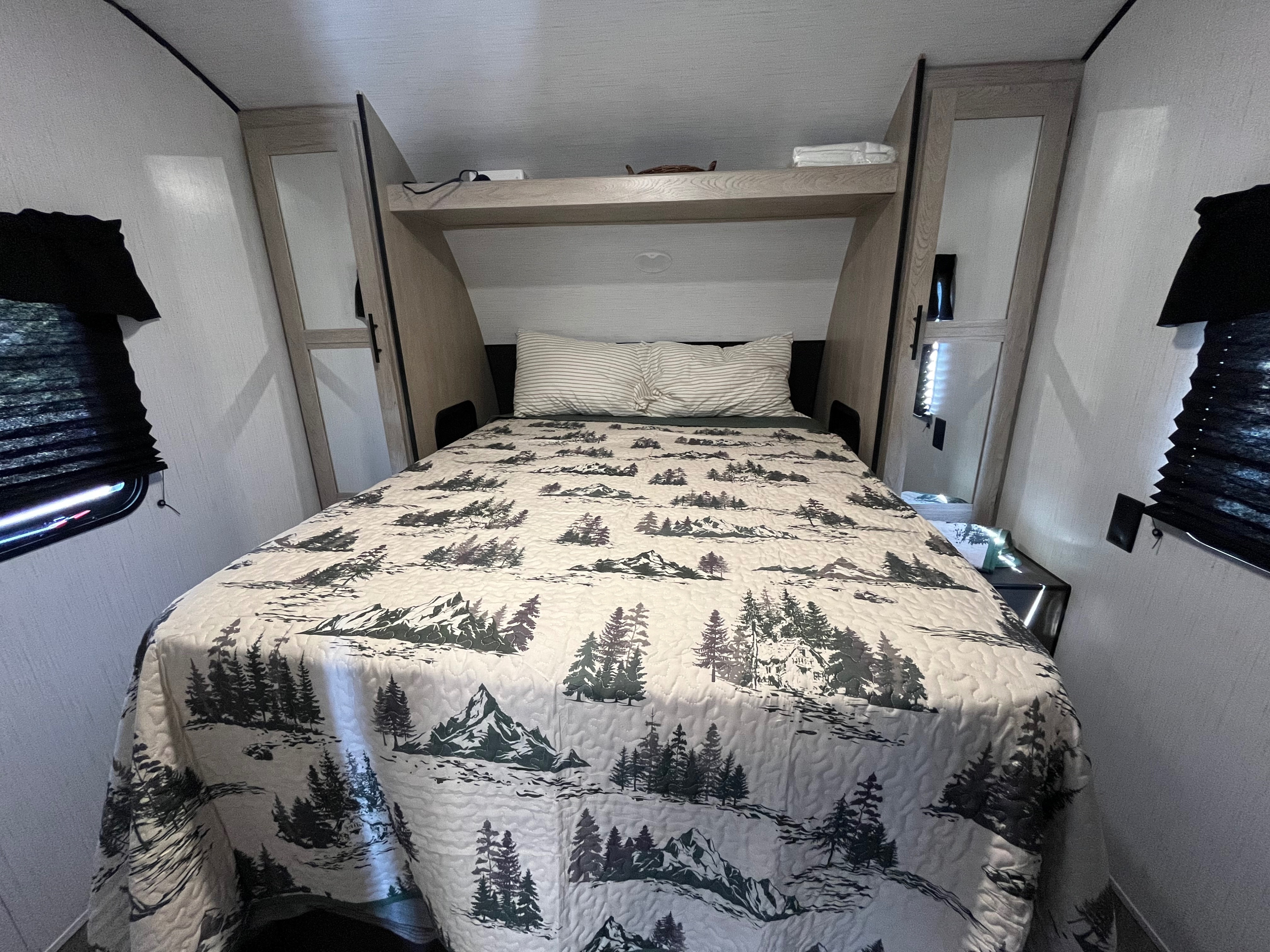 Cozy rv rental bedroom featuring a nature-themed quilt, striped pillows, built-in storage, and side windows with blinds.. Heartland RVs Prowler 2021
