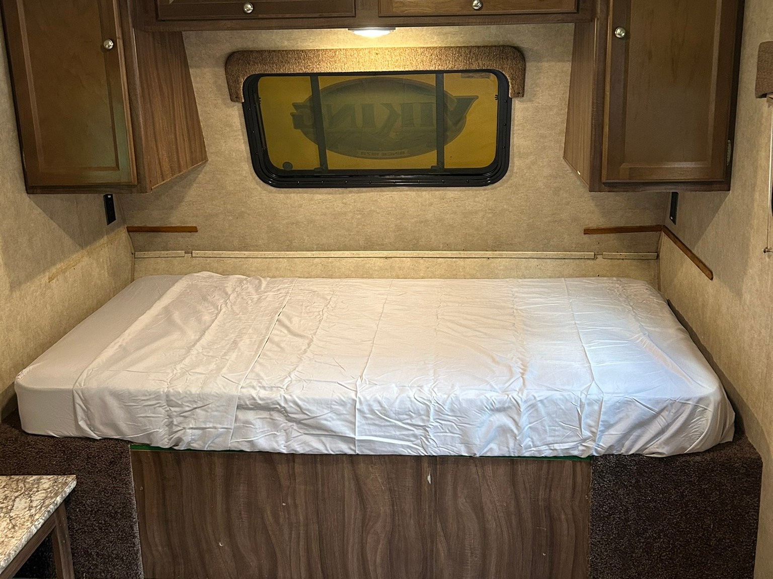 Cozy RV rental bedroom interior, featuring a neatly made bed with white sheets, wood cabinets, and a window.. Forest River Viking 2021