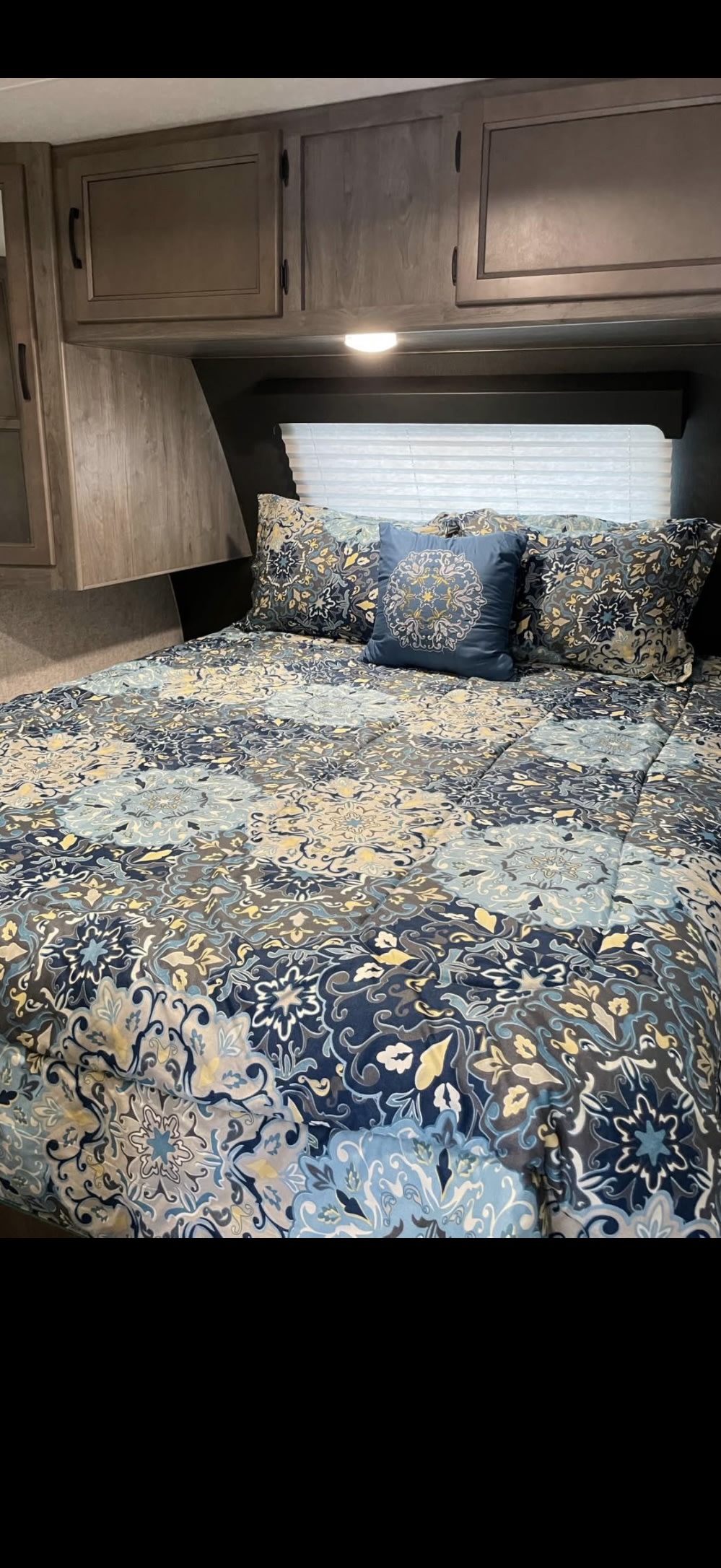 Cozy RV rental bedroom featuring patterned blue and yellow bedding, overhead wood cabinets, and window blinds.. Coachmen Apex 2021