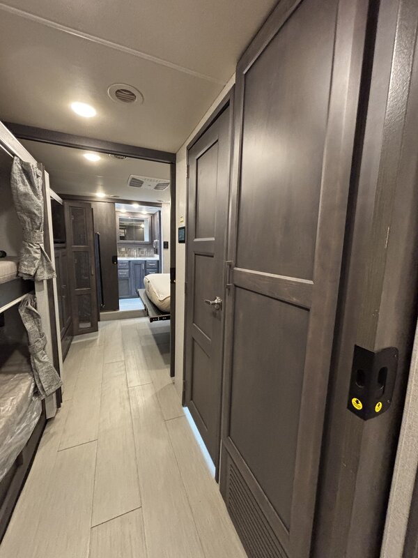An RV rental interior: modern hallway with light wood-look floor, bunk beds (left), dark cabinets (right), leading to the kitchen/bathroom.. Thor Palazzo 2023