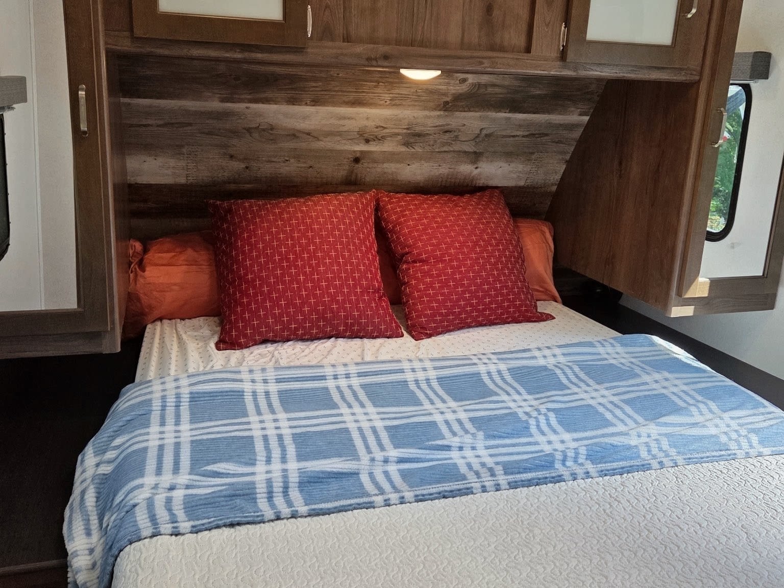 Cozy RV rental bedroom featuring a wooden headboard, red pillows, and a blue plaid blanket. Relaxing space for your travel adventure.. Keystone RV Passport Grand Touring 2020