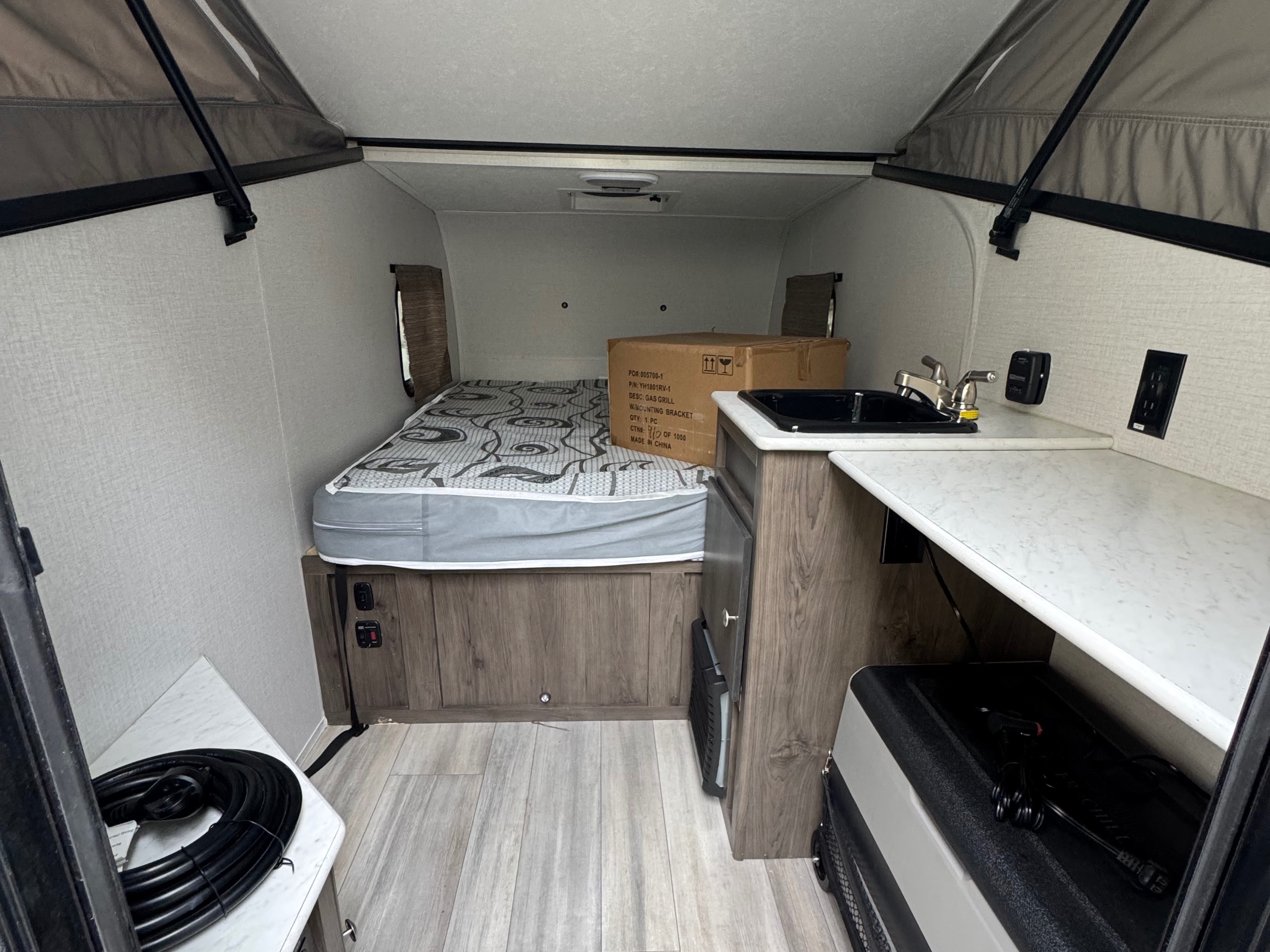 Inside a compact RV rental: bed, small kitchen with sink, a box (gas grill), power cord, and pop-top roof for camping.. Forest River Coachmen Clipper 2023