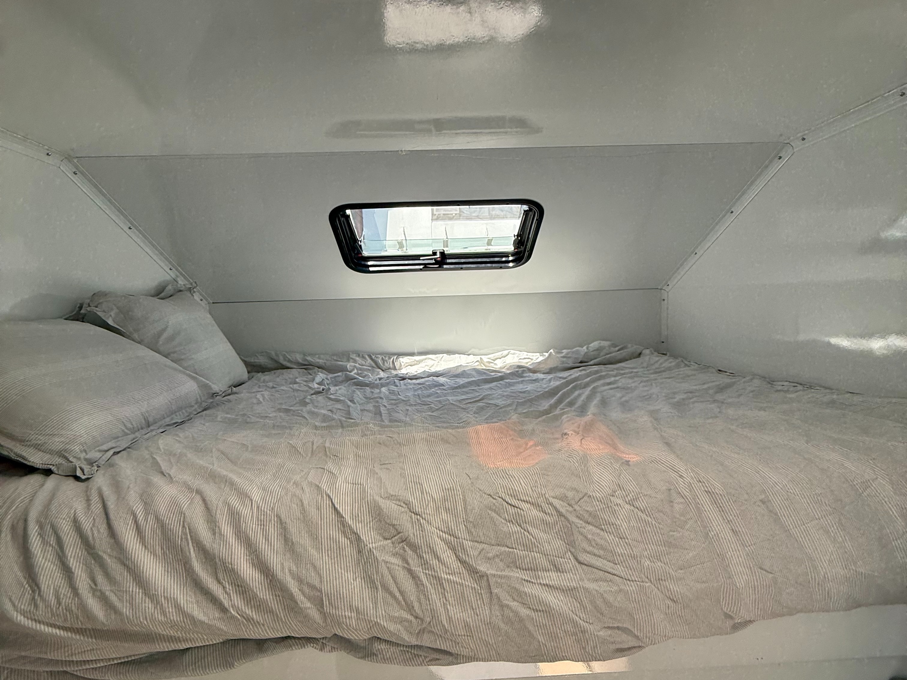 Cozy RV rental bedroom featuring a striped bed, pillows, and an overhead window. Compact and comfortable sleeping area inside.. Custom Camper 2025