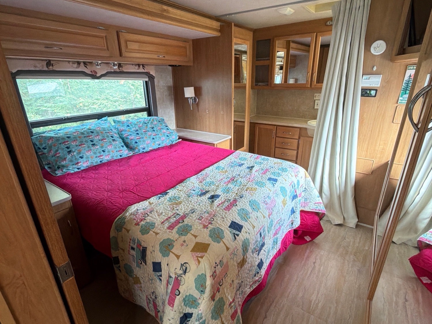 Cozy RV rental bedroom featuring a colorful bed, large window with greenery, and warm wood cabinetry.. Fleetwood Bounder 2008