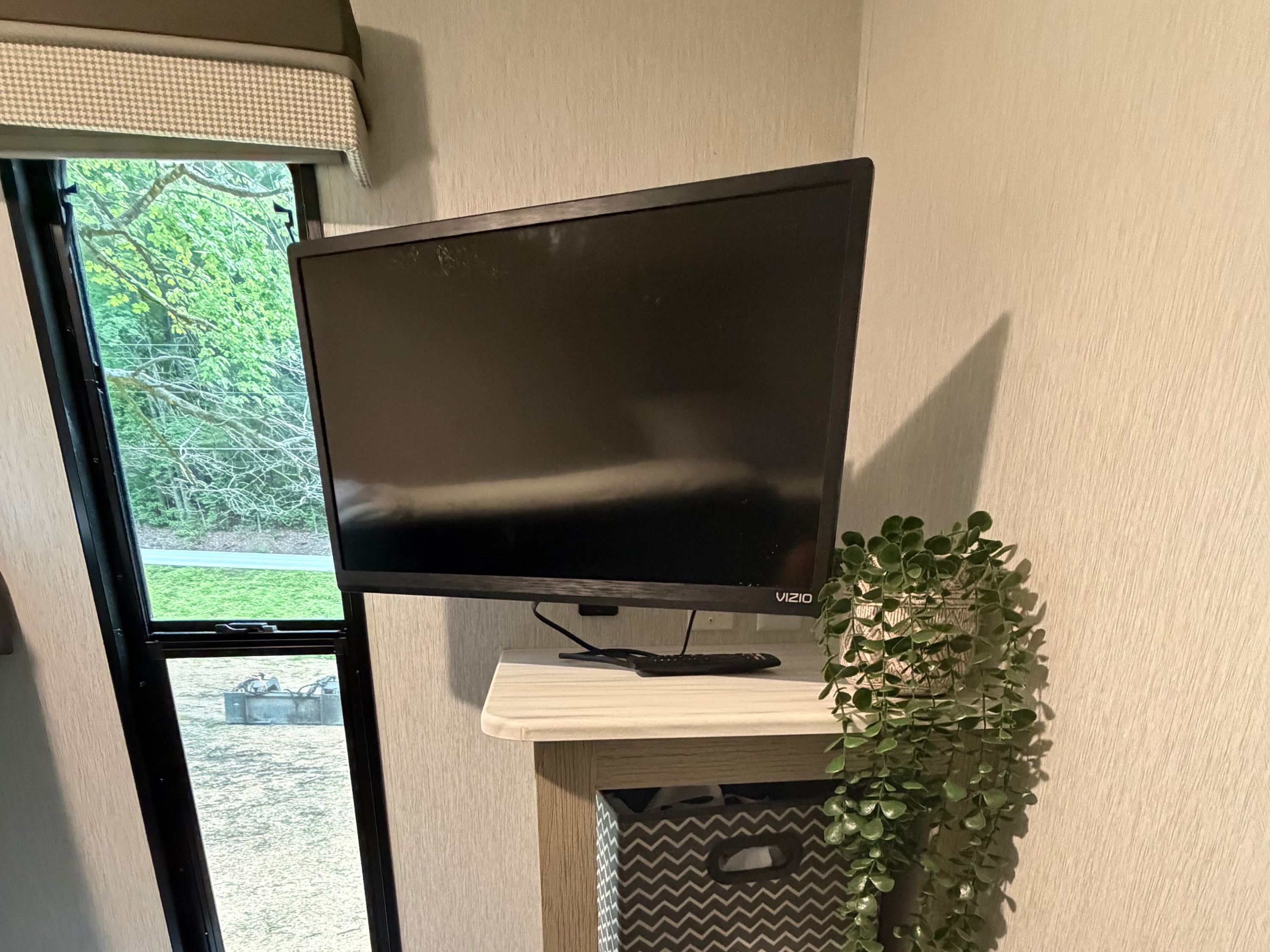 Cozy rv rental interior: TV over shelves with storage bins & a plant, beside a window with green trees outside.. Forest River Salem Cruise Lite Platinum 2023
