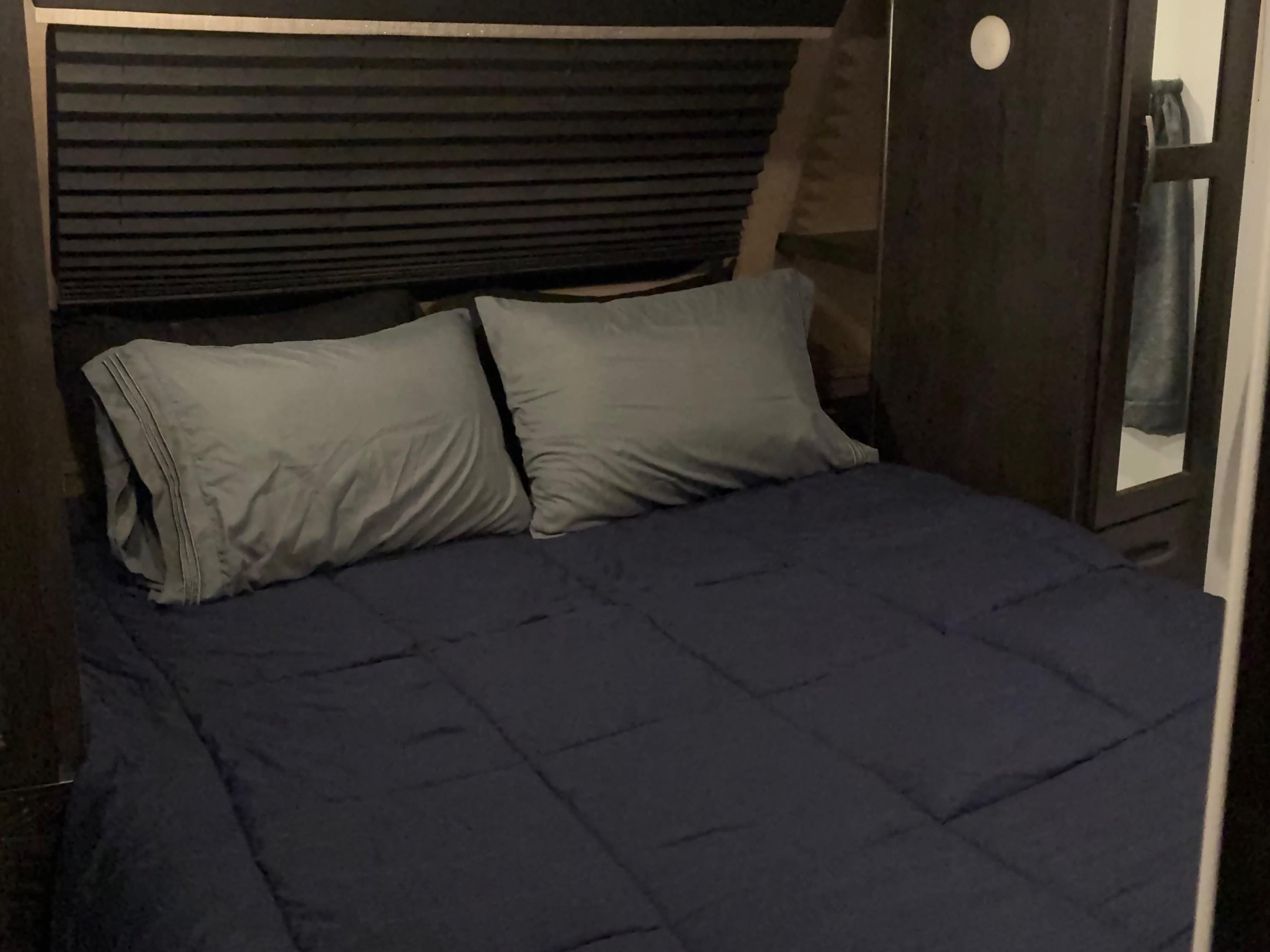 Dark RV rental bedroom with a bed, grey pillows, navy comforter, pleated blind, and built-in cabinet with mirror.. Forest River Surveyor Luxury 2019